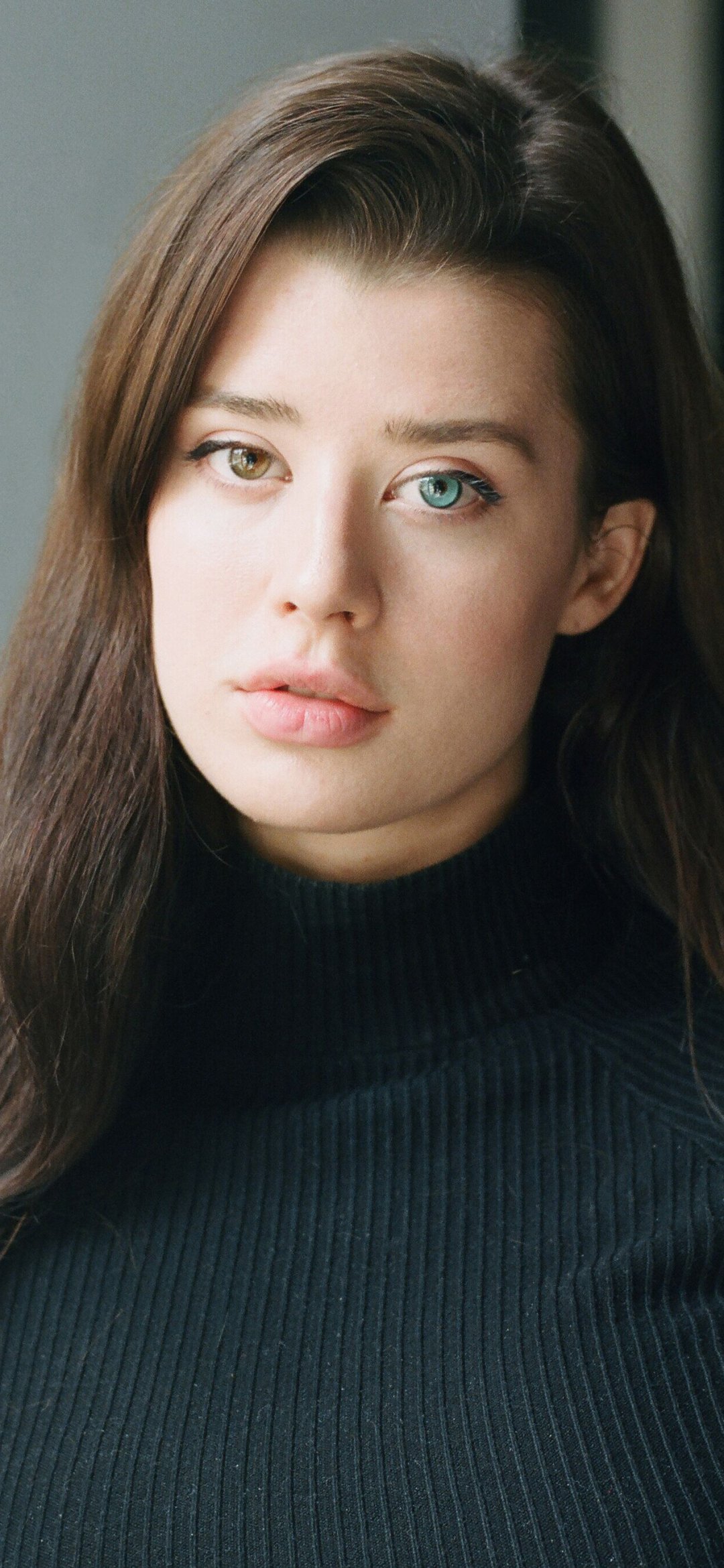 Phone wallpaper: close-up of a woman model with long brown hair, green and blue eyes, neutral expression, wearing a black turtleneck in soft natural light.
