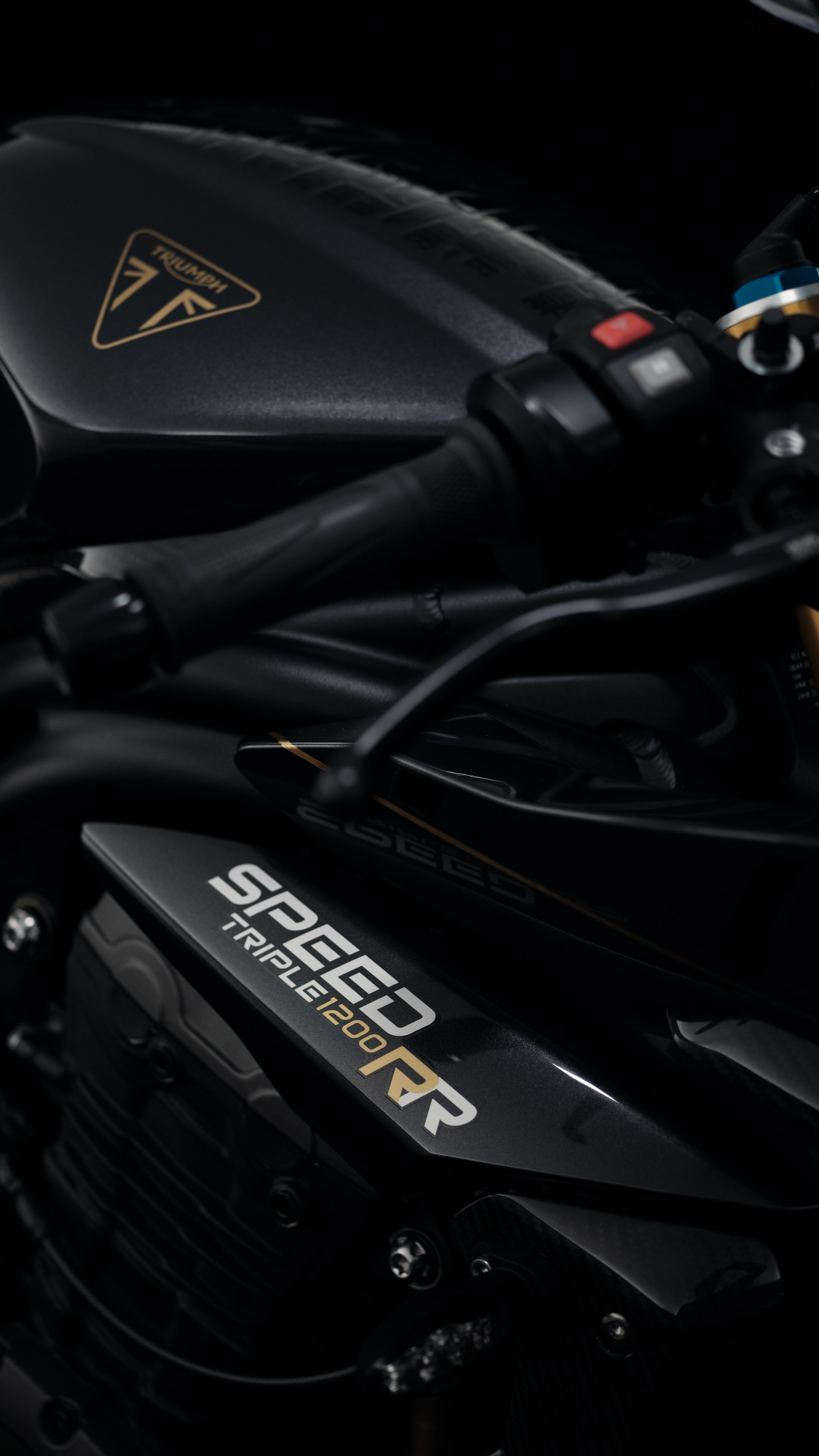 Close-up of the Triumph Speed Triple 1200 RR Bond Edition, showcasing sleek black details and the iconic logo, set against a dark background for a striking phone wallpaper.