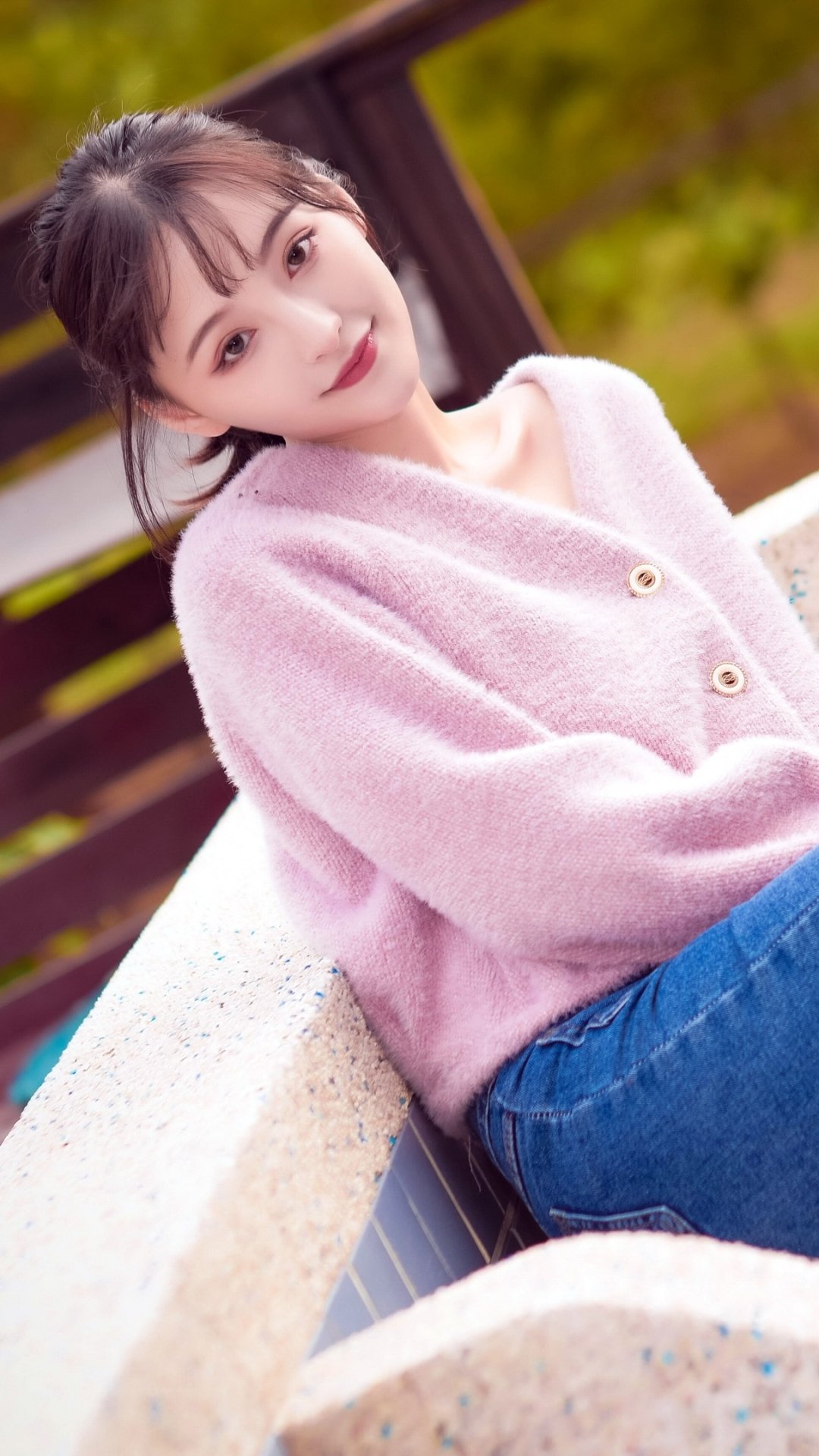 Phone wallpaper featuring a young Asian woman in a soft pink sweater leaning on a stone bench, with a blurred green and wooden background for iPhone and Android screens.