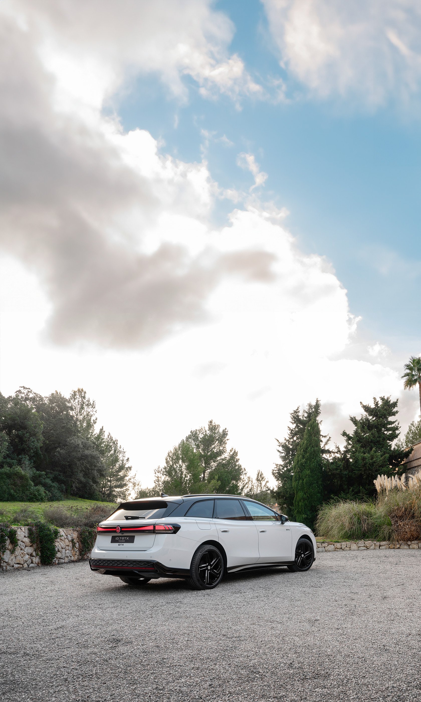 A sleek Volkswagen ID.7 GTX Tourer parked in a scenic outdoor setting, surrounded by greenery under a partly cloudy sky. This image makes an appealing phone wallpaper.