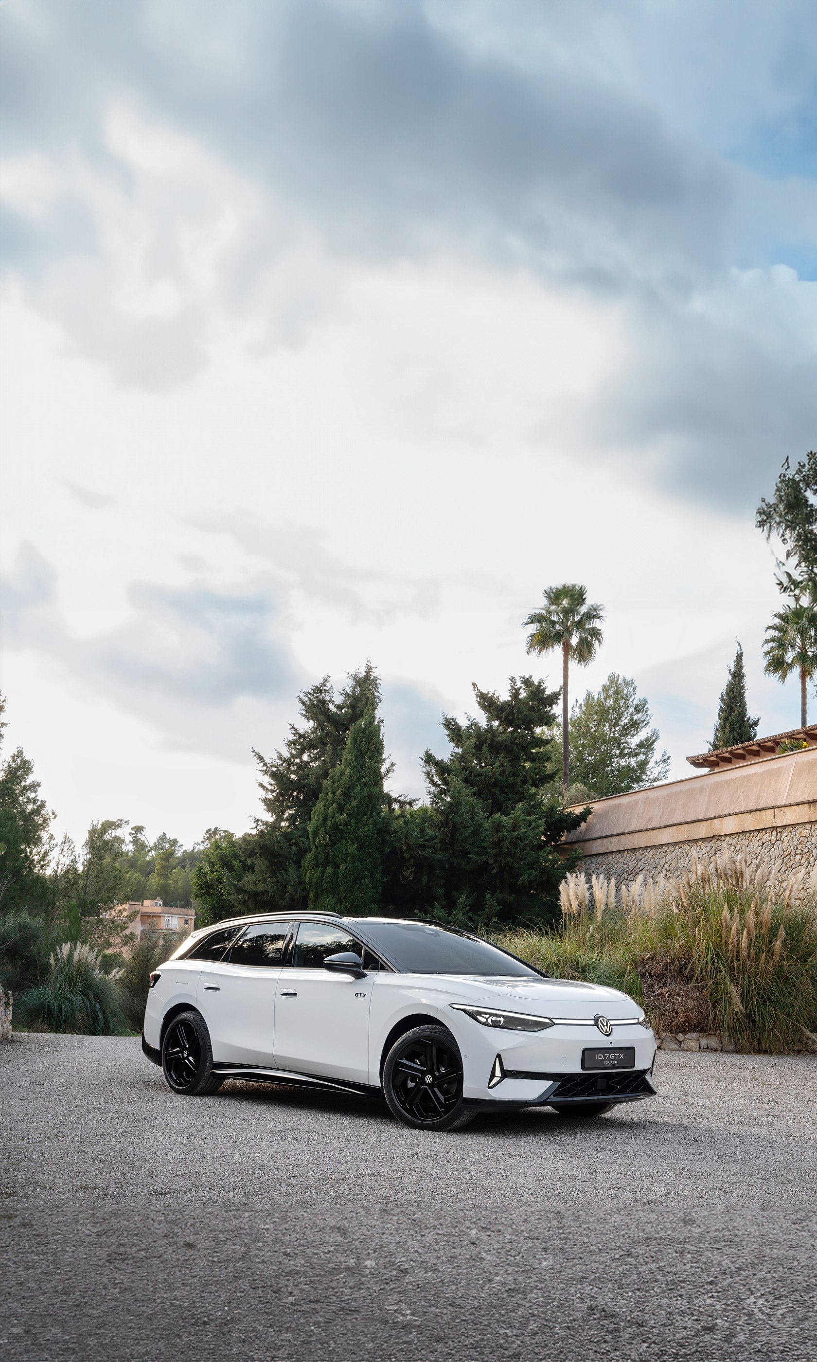A sleek Volkswagen ID.7 GTX Tourer parked in a serene setting, surrounded by greenery and palm trees, with a dramatic sky in the background, designed as a striking phone wallpaper.