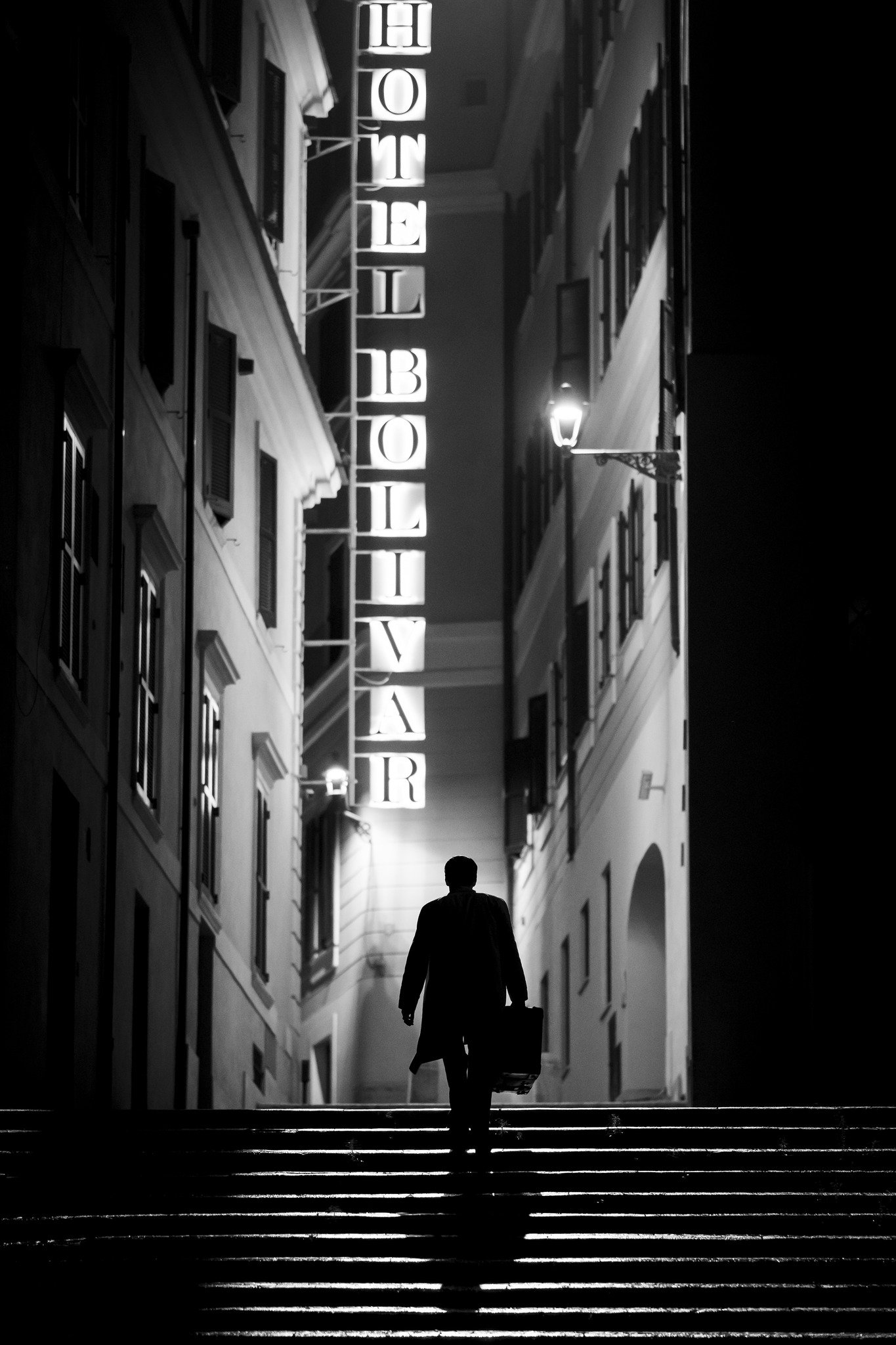 Black and white phone wallpaper featuring a silhouette of a person walking towards the Hotel Bolivar sign in an atmospheric alleyway, evoking the mood of the TV show Ripley.