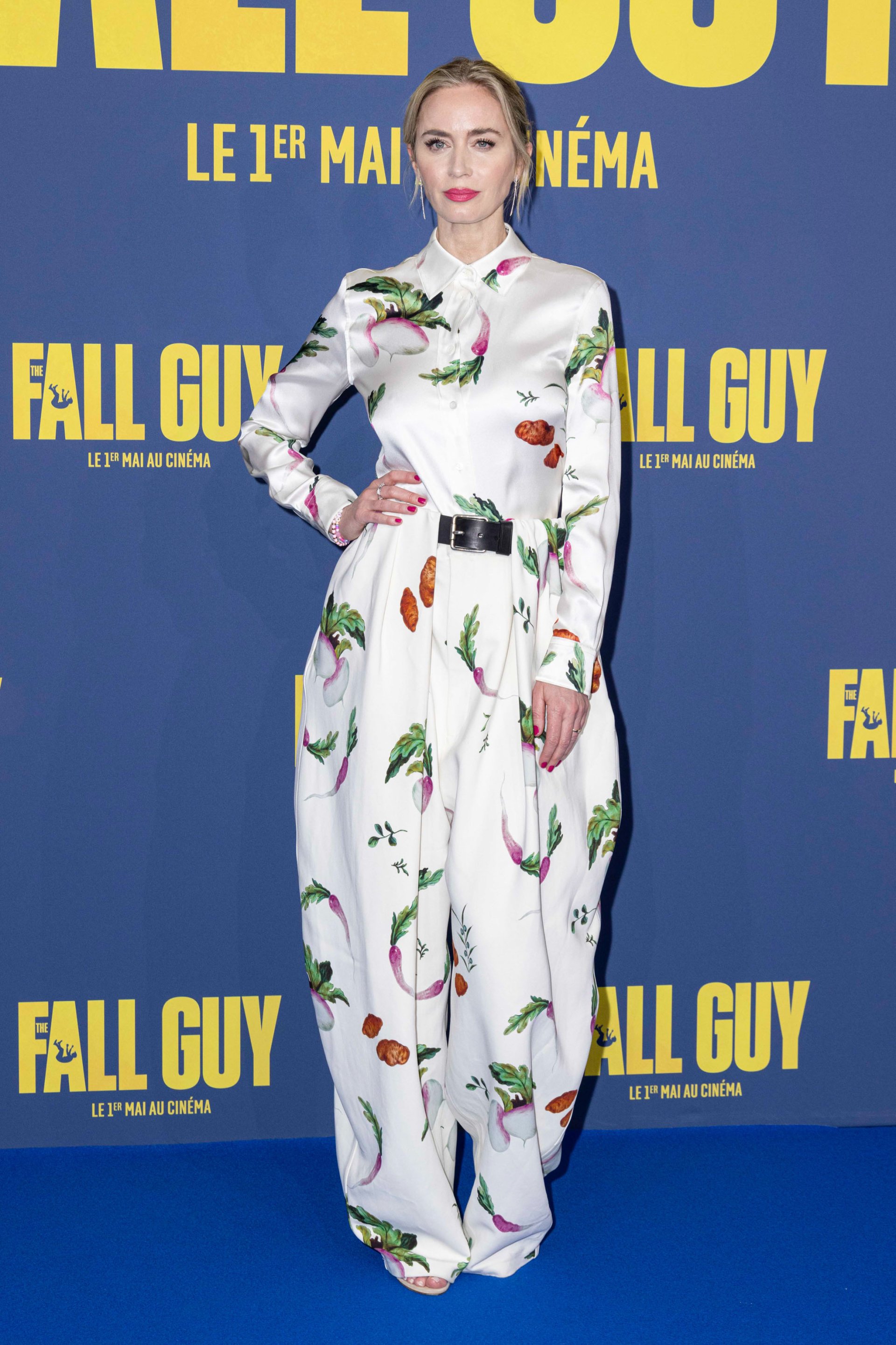 Emily Blunt in a fruit-patterned dress at 'The Fall Guy' movie premiere, posing against a blue backdrop with the movie title. Good as a phone wallpaper.