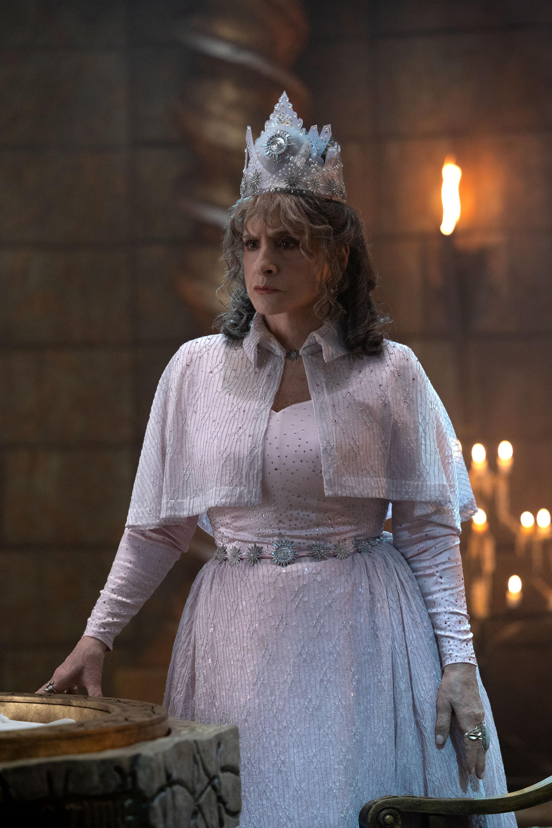 A scene from the TV show Agatha All Along, featuring a character in a regal outfit and crown, standing in a dimly lit room with candles, creating an intriguing atmosphere.