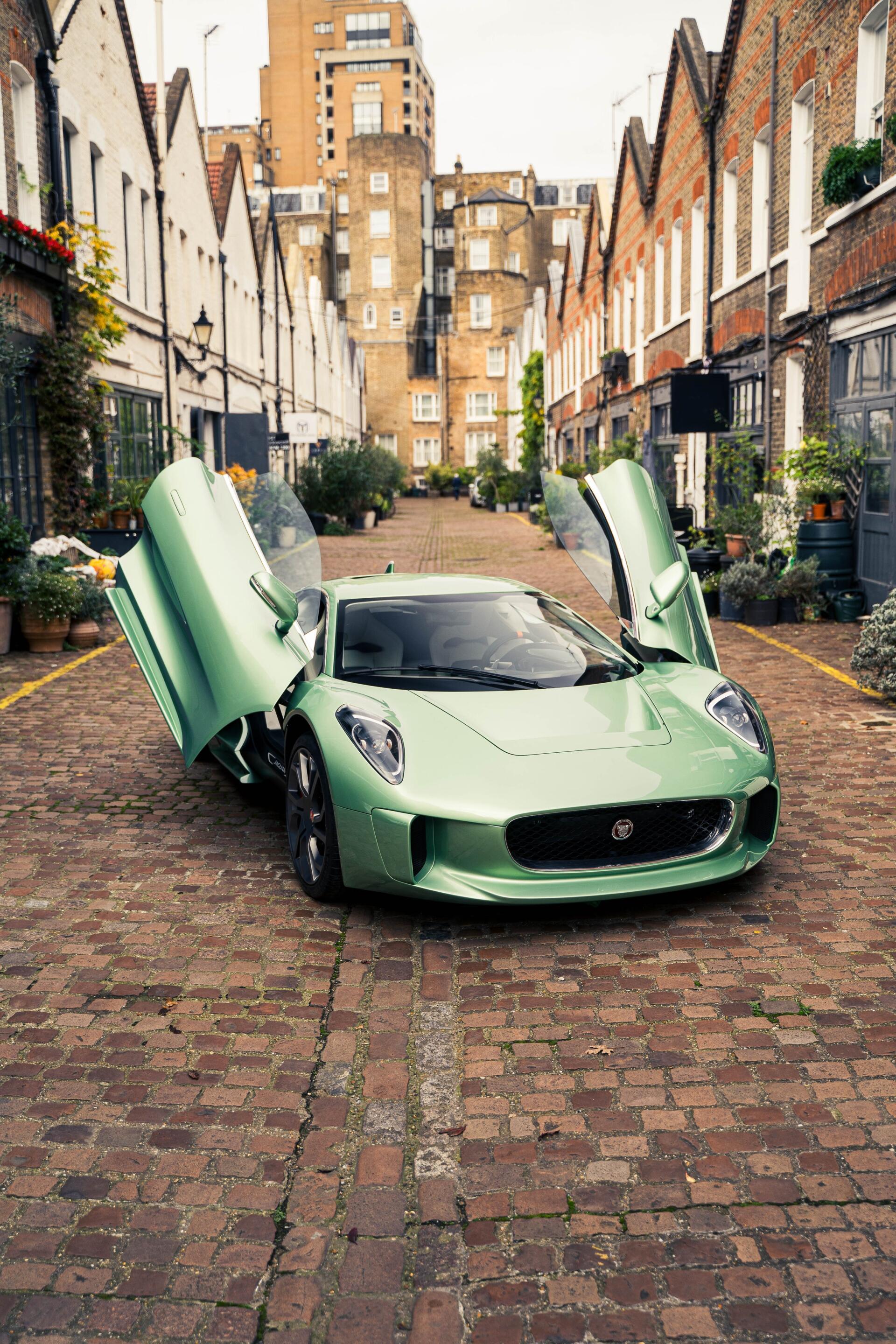 A stunning Jaguar C-X75 in a vibrant green color, parked on a cobblestone street with its doors raised, showcasing its luxury design amid charming urban architecture.
