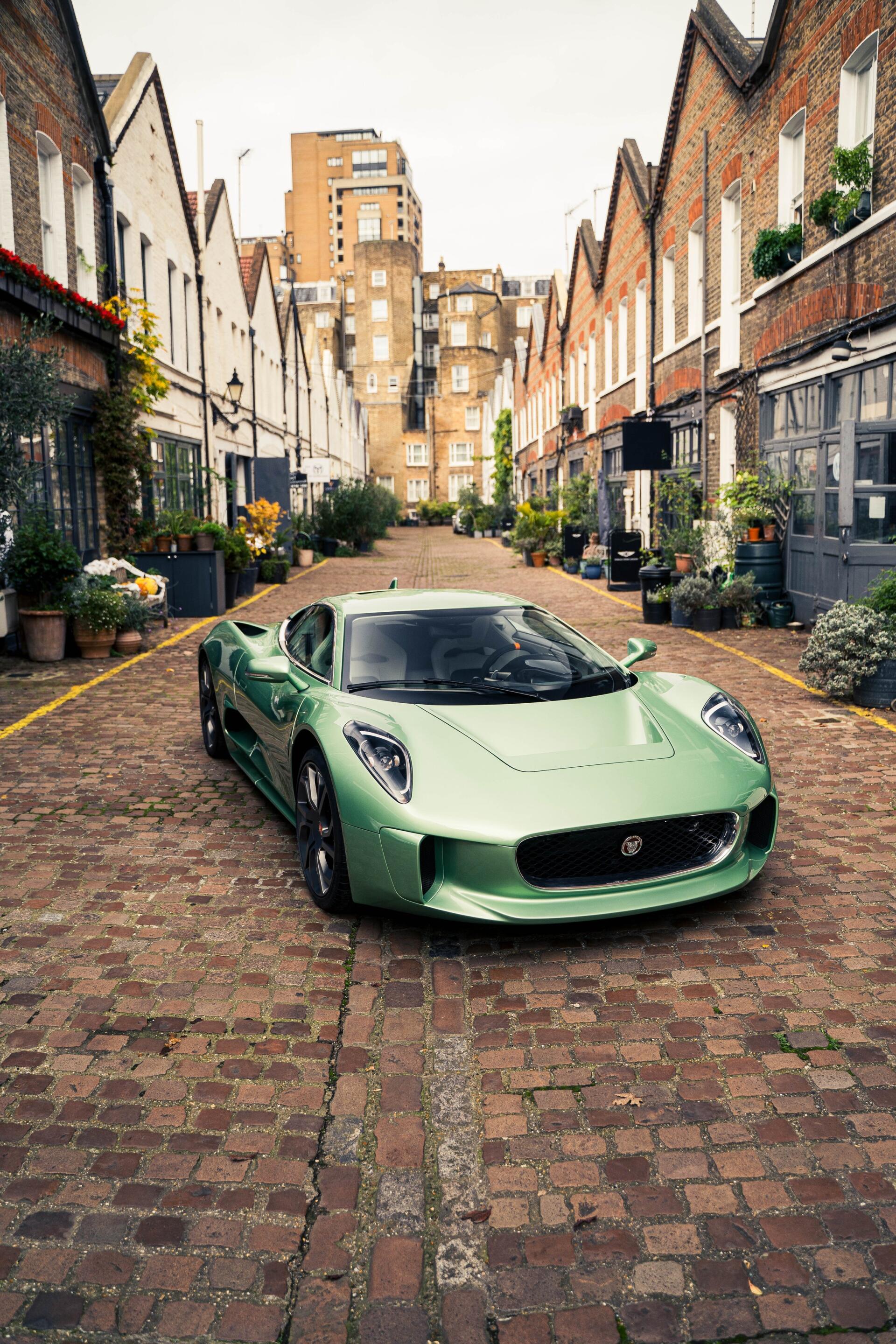 A sleek, green Jaguar C-X75 sports car is parked in a charming cobblestone alley lined with quaint buildings and gardens, creating a striking phone wallpaper.