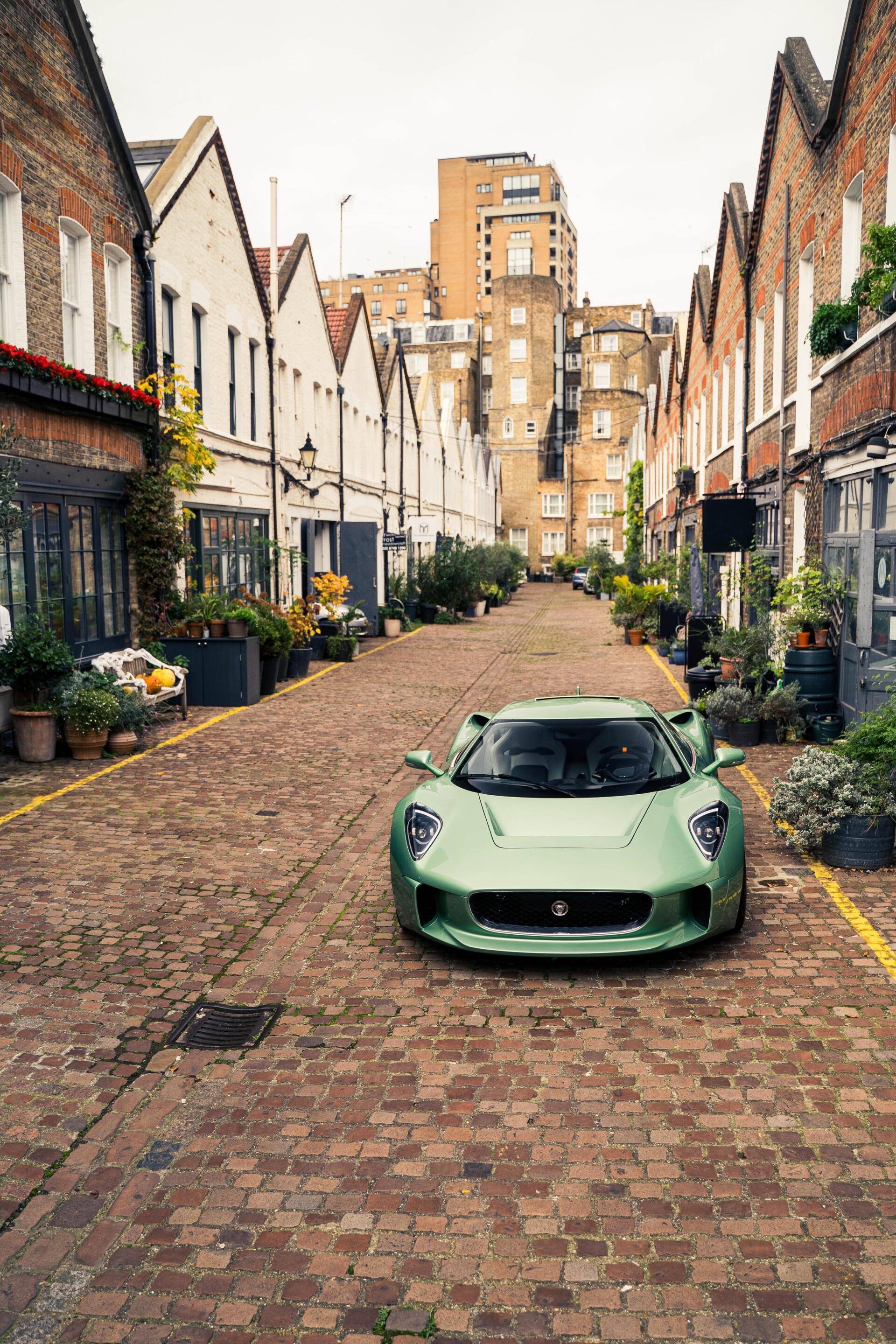A sleek green Jaguar C-X75 parked in a charming cobblestone alley, framed by quaint buildings and vibrant greenery, making for an eye-catching phone wallpaper.