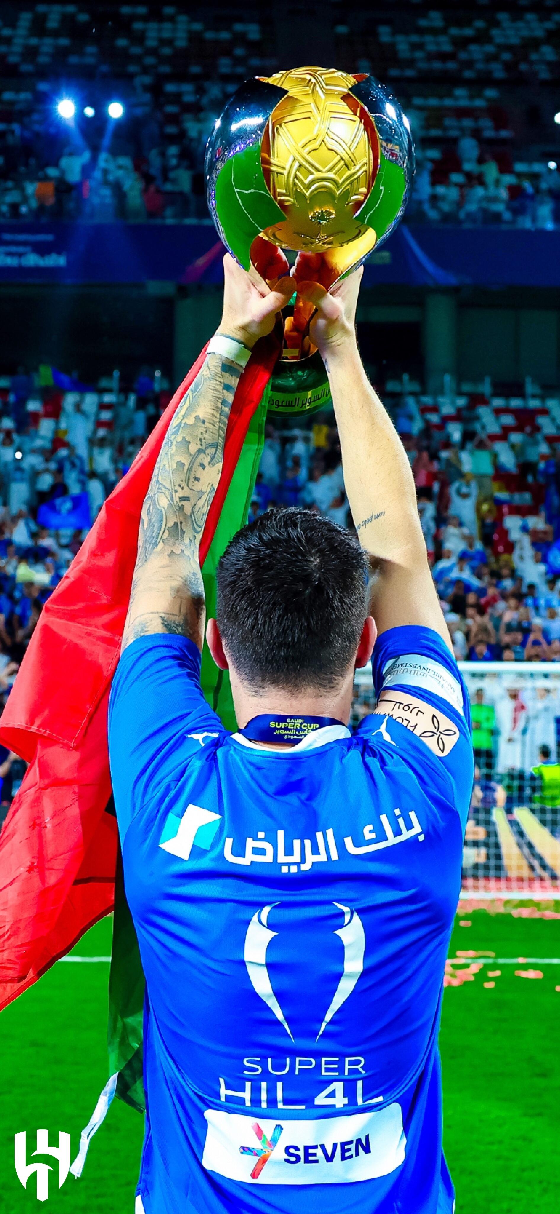 A jubilant player from Al Hilal SFC holds a championship trophy high, draped in a vibrant flag, celebrating a triumphant moment on the field, with a cheering crowd in the background.
