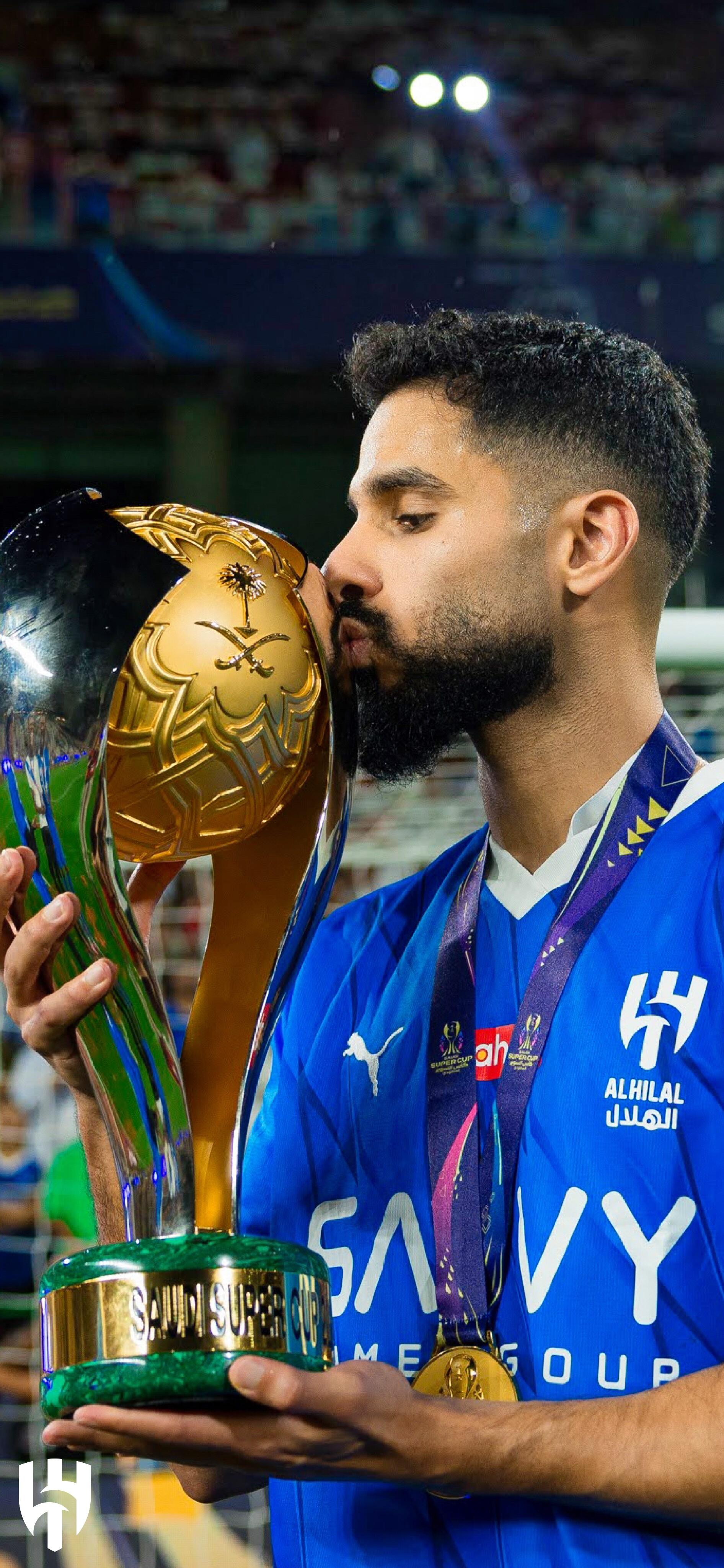 A player from Al Hilal SFC kisses their trophy, celebrating a victory. The athlete wears a blue jersey and a medal, capturing a triumphant moment in sports.