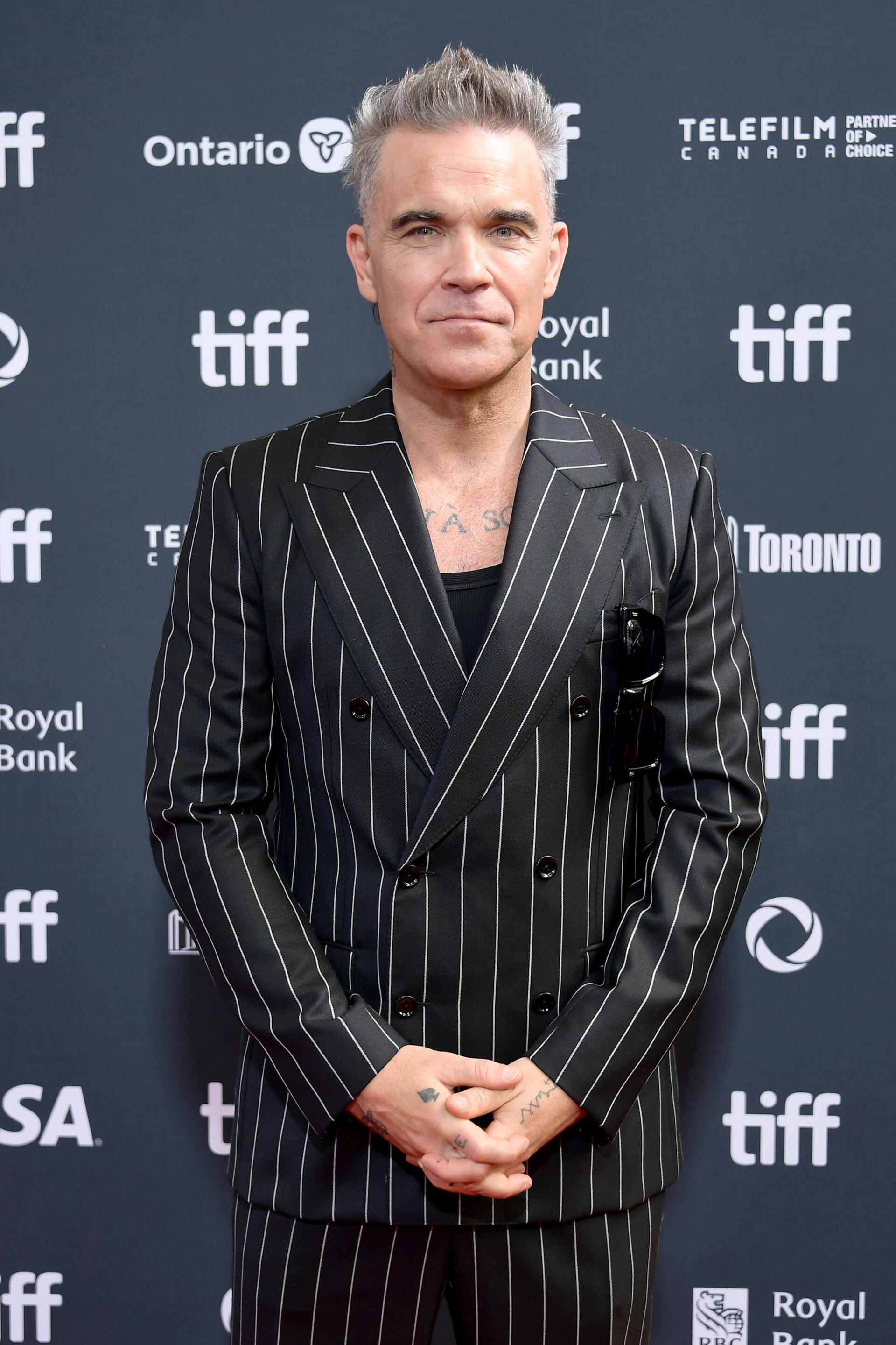 Robbie Williams poses in a stylish pinstripe suit at the premiere of Better Man (2024) during TIFF, showcasing his celebrity status against a branded backdrop.