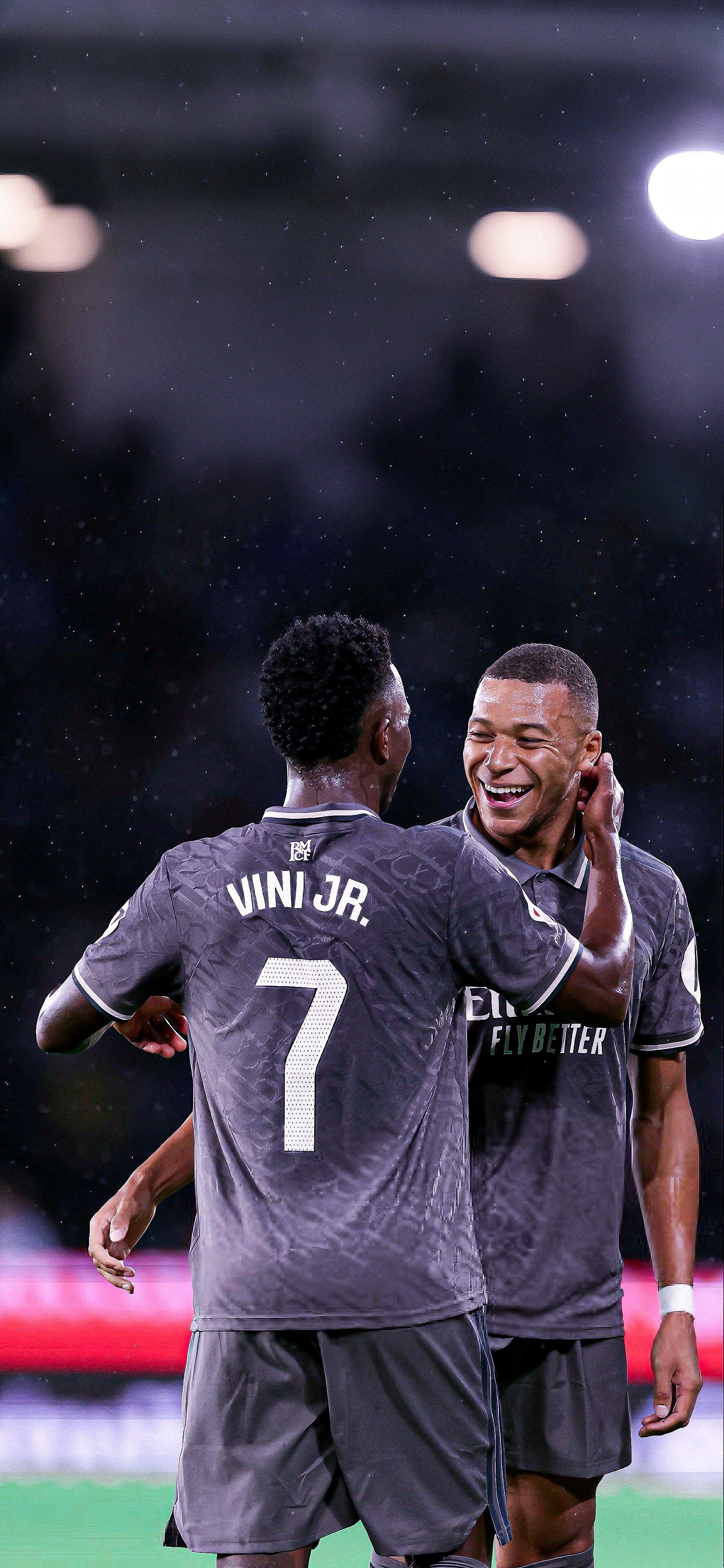 Kylian Mbappé and Vinícius Júnior share a joyful moment on the field, both dressed in Real Madrid C.F. jerseys, celebrating a play amidst a dynamic soccer match.