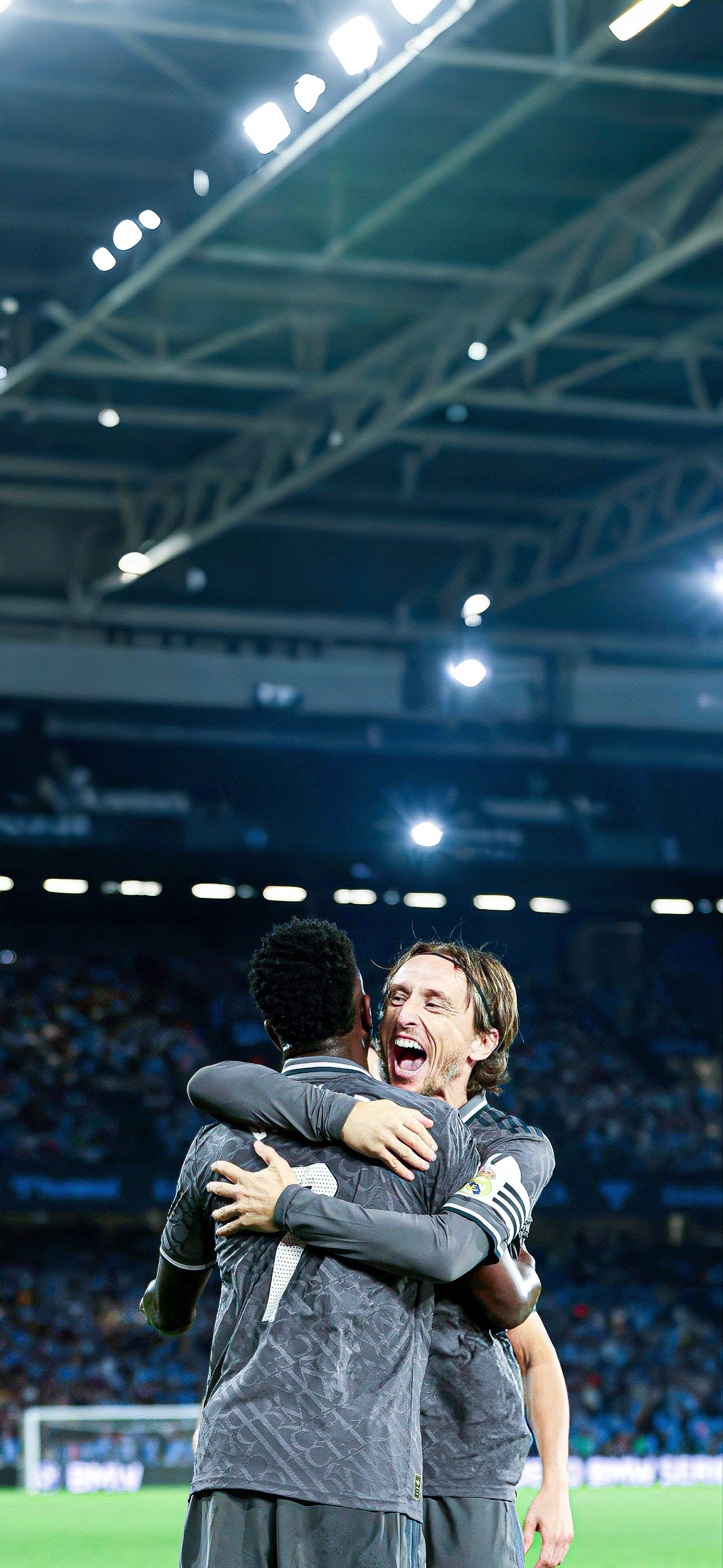 Luka Modrić celebrates passionately with a teammate during a Real Madrid C.F. match, surrounded by an energetic crowd under bright stadium lights. A striking soccer phone wallpaper.