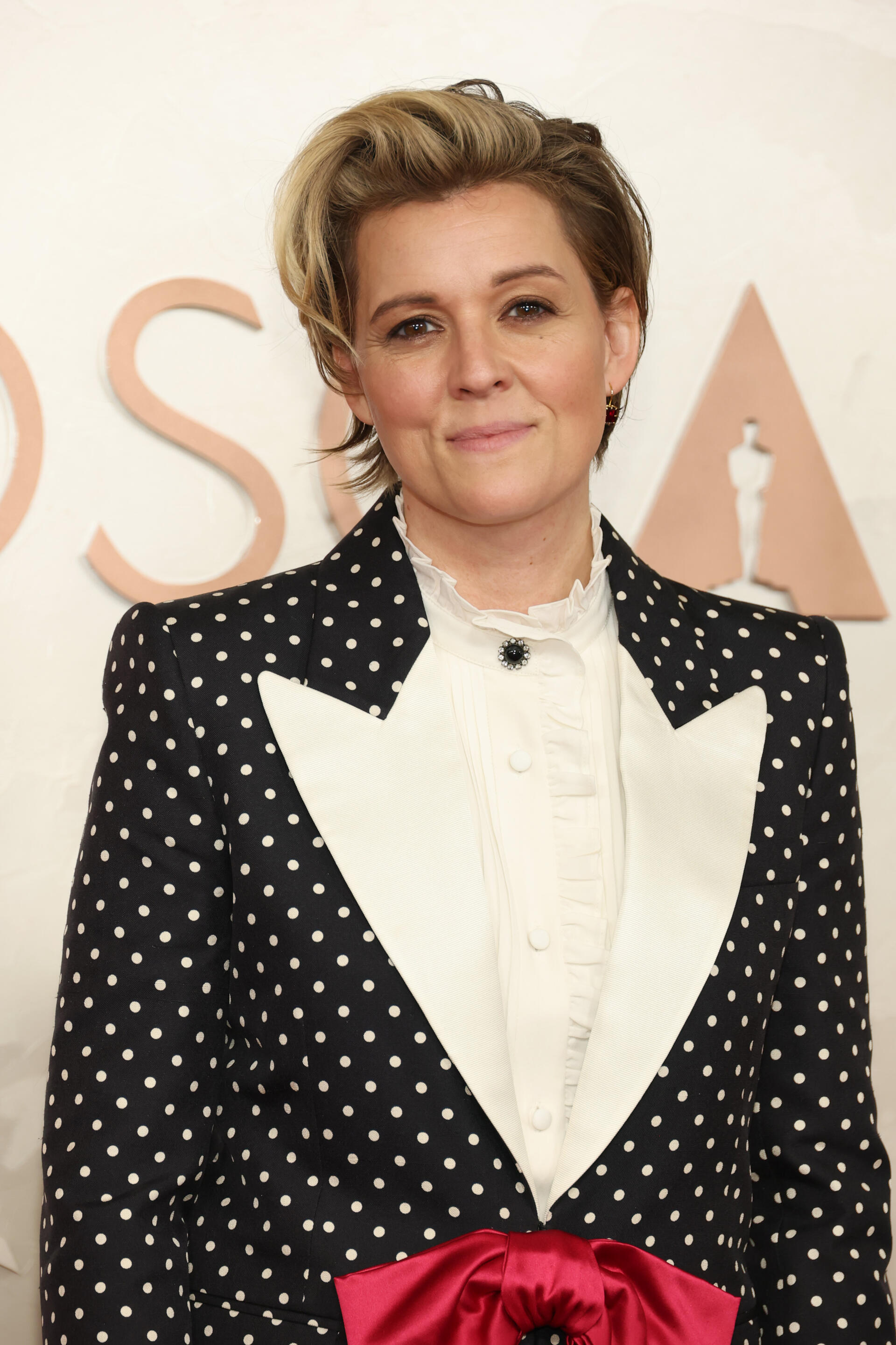 Brandi Carlile poses at the Academy Awards, wearing a stylish polka-dot tuxedo with a white blouse and a red bow, showcasing a blend of elegance and modern flair.