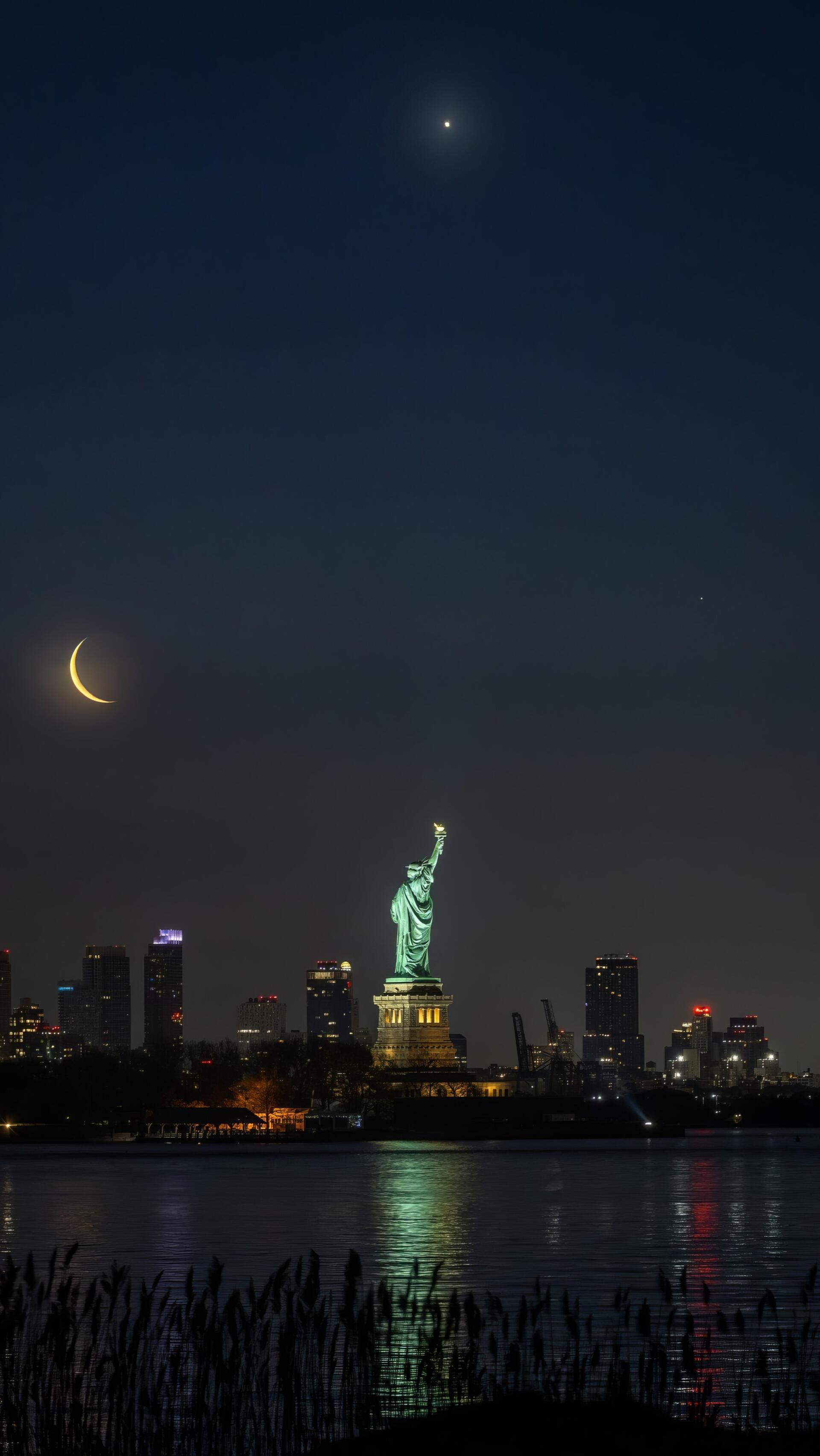 Night view of the illuminated Statue of Liberty in New York with a crescent moon, captured as a vertical phone wallpaper compatible with iPhones and Android devices.