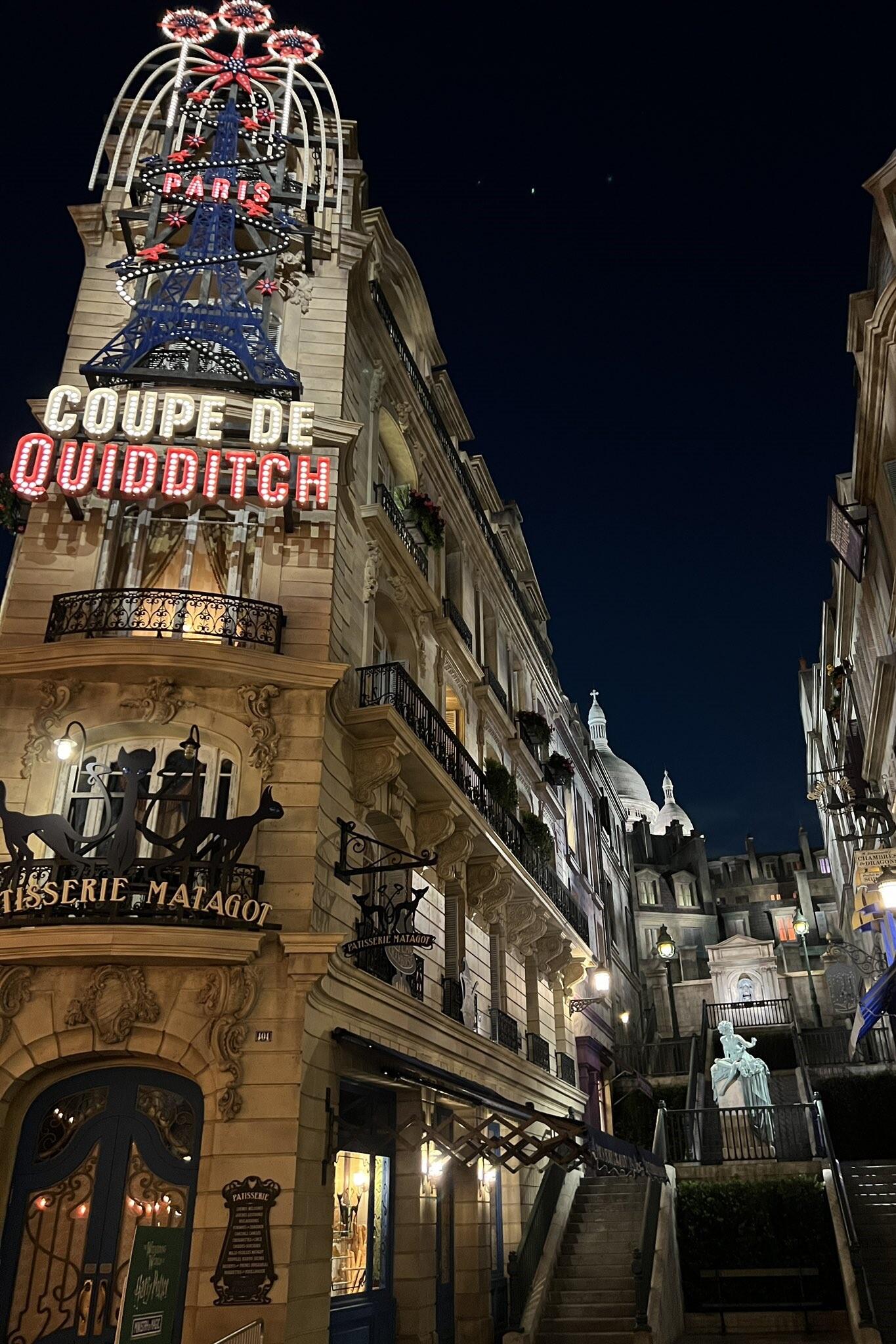 Nighttime view of the Paris-themed Universal Studios building with a glowing Eiffel Tower sign and Coupe de Quidditch marquee, captured as a phone wallpaper for iPhones and Android.