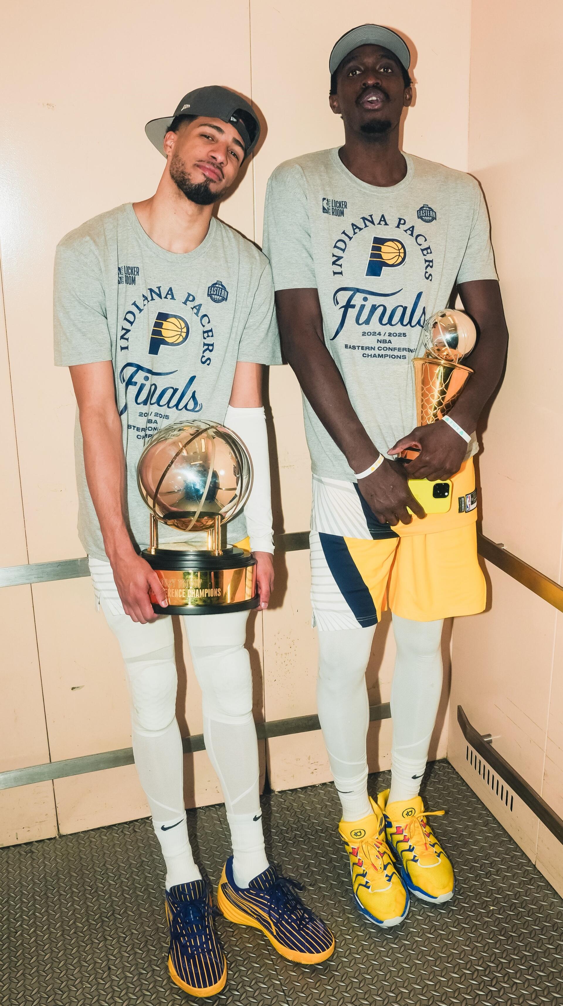 Pascal Siakam and Tyrese Haliburton of the Indiana Pacers hold NBA championship trophies, wearing matching Finals shirts in a celebratory basketball moment.