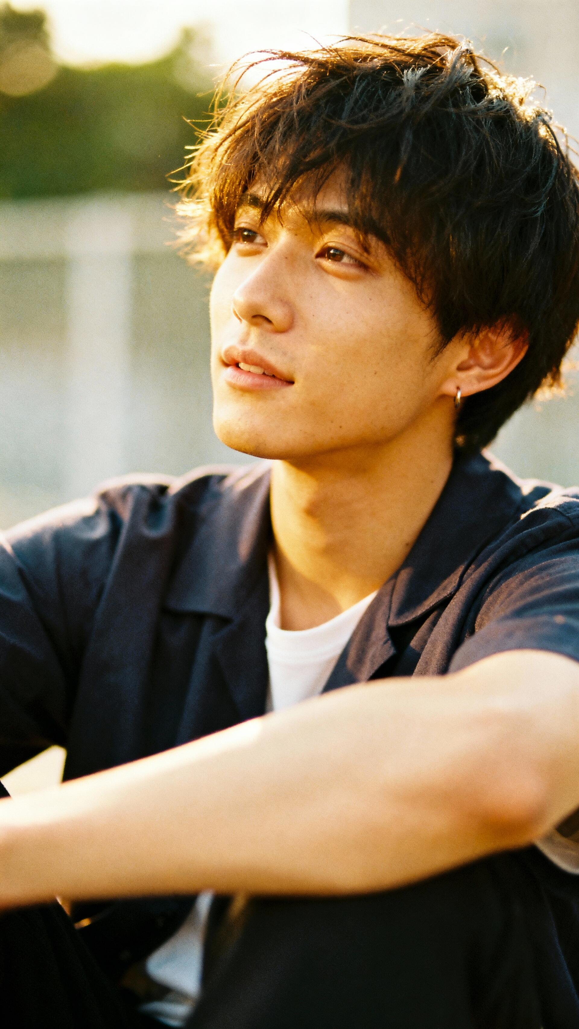Phone wallpaper showing a young man with tousled hair and an earring, seated outdoors at golden hour, gazing upward with a gentle smile and warm sunlight on his face.