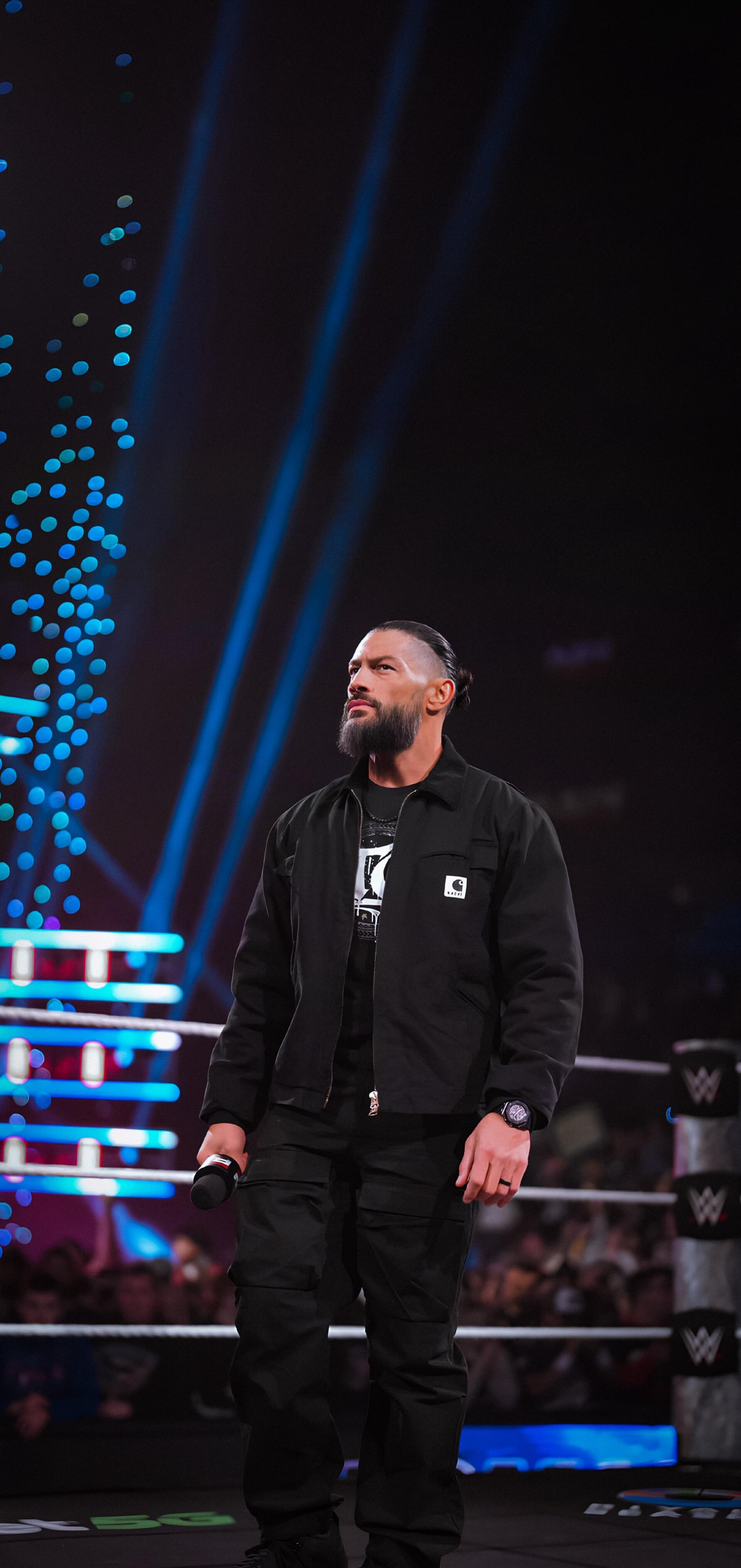 Vertical phone wallpaper of a lone figure standing in a WWE ring holding a microphone under blue spotlights, with the crowd, ring ropes, and WWE branding visible in the background.