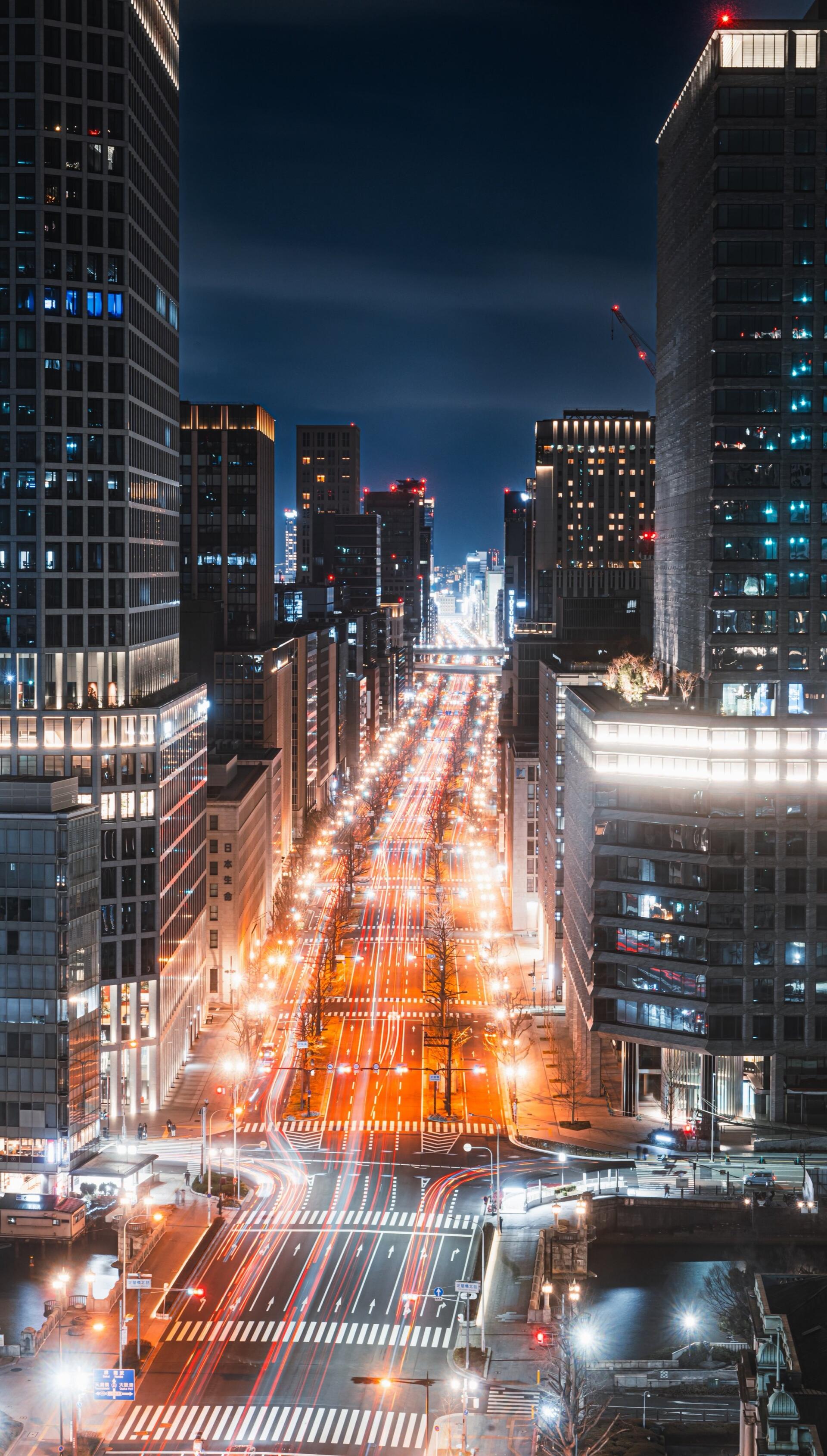 Vertical phone wallpaper of a Japanese cityscape at night: illuminated downtown avenue lined with towering buildings, long light trails and a glowing street grid.