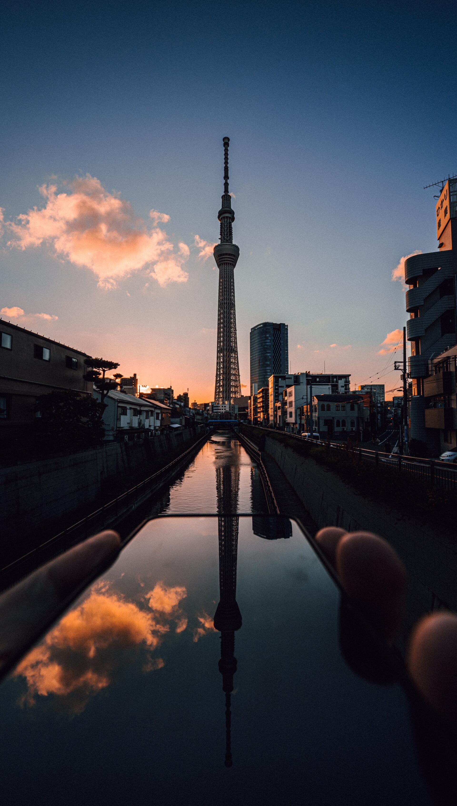 Vertical phone wallpaper of Tokyo Skytree towering over a Tokyo canal at sunset, its silhouette reflected in calm water with city buildings lining the banks.