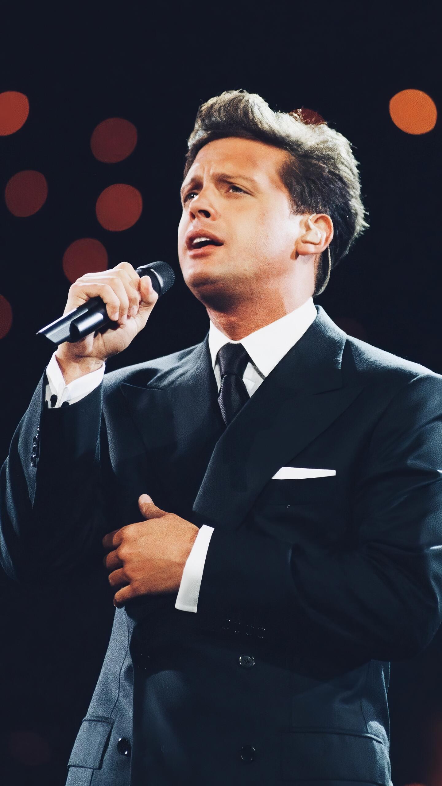 Phone wallpaper showing a male singer in a dark suit, holding a microphone and performing on stage with warm bokeh lights behind him.