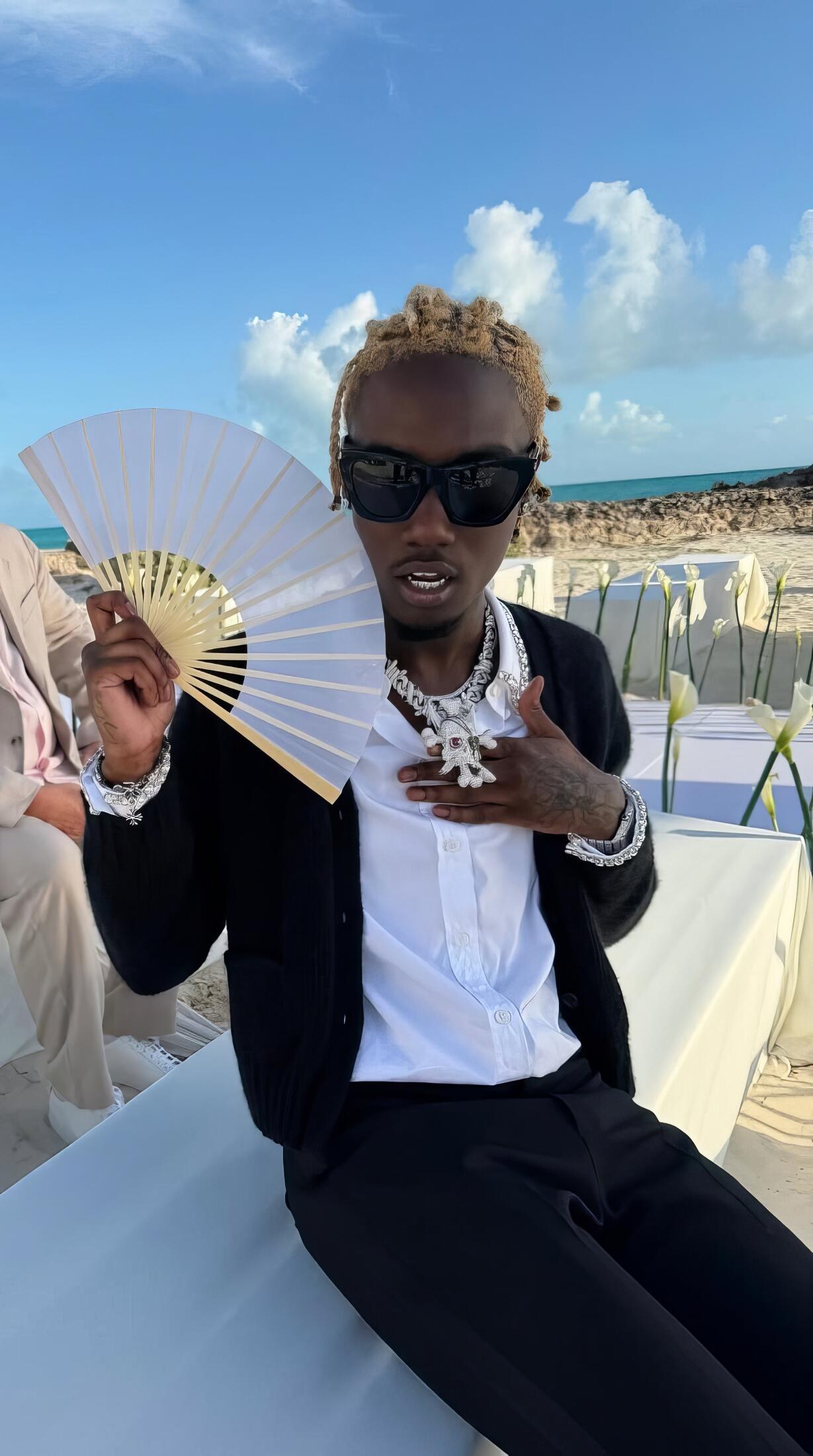 Phone wallpaper of a rapper flexing sunglasses and heavy jewelry on a beach, holding a white folding fan and posing confidently.