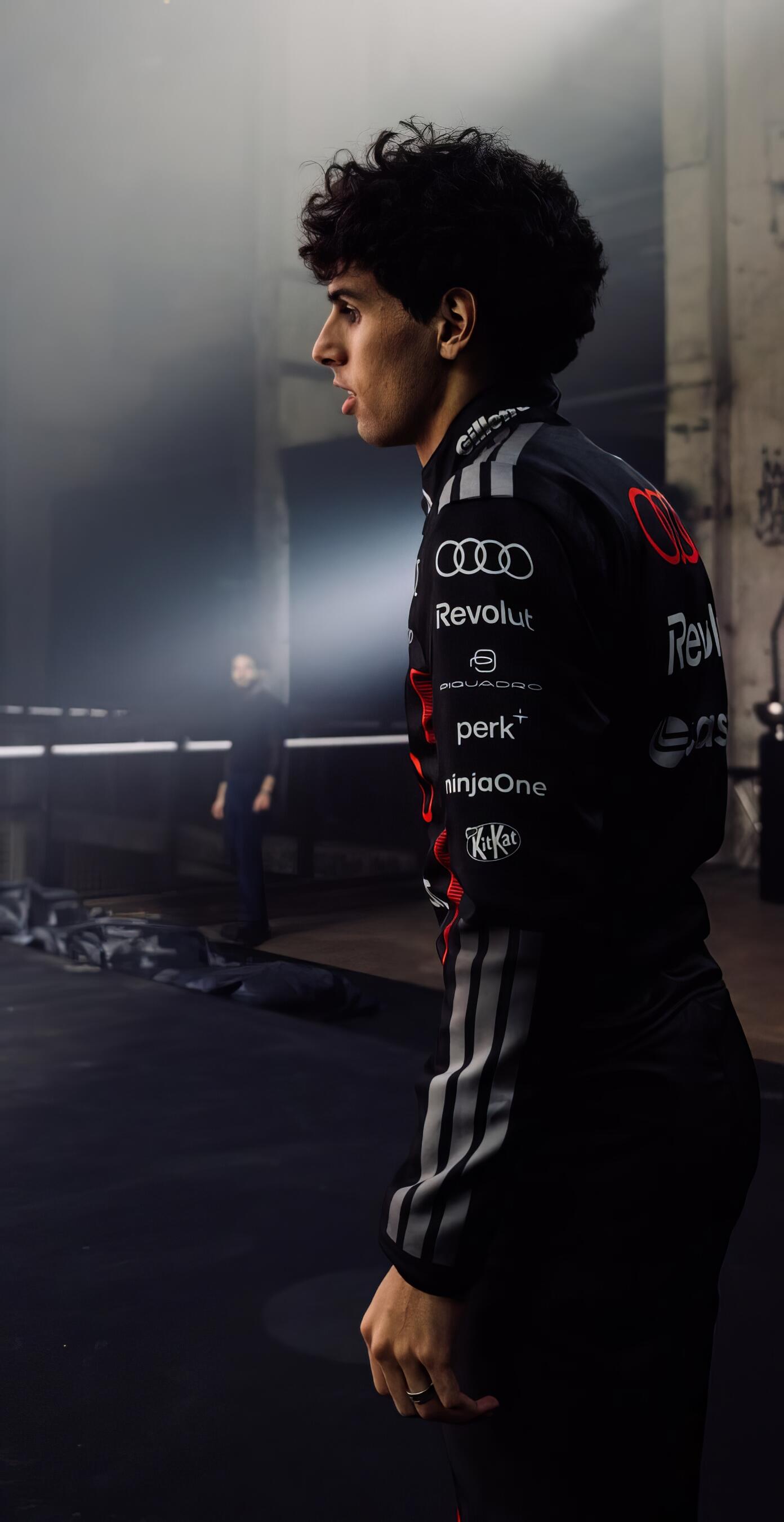 Phone wallpaper: profile of a young man in an Audi F1 racing suit standing in a dim garage, dramatic side lighting and moody atmosphere.