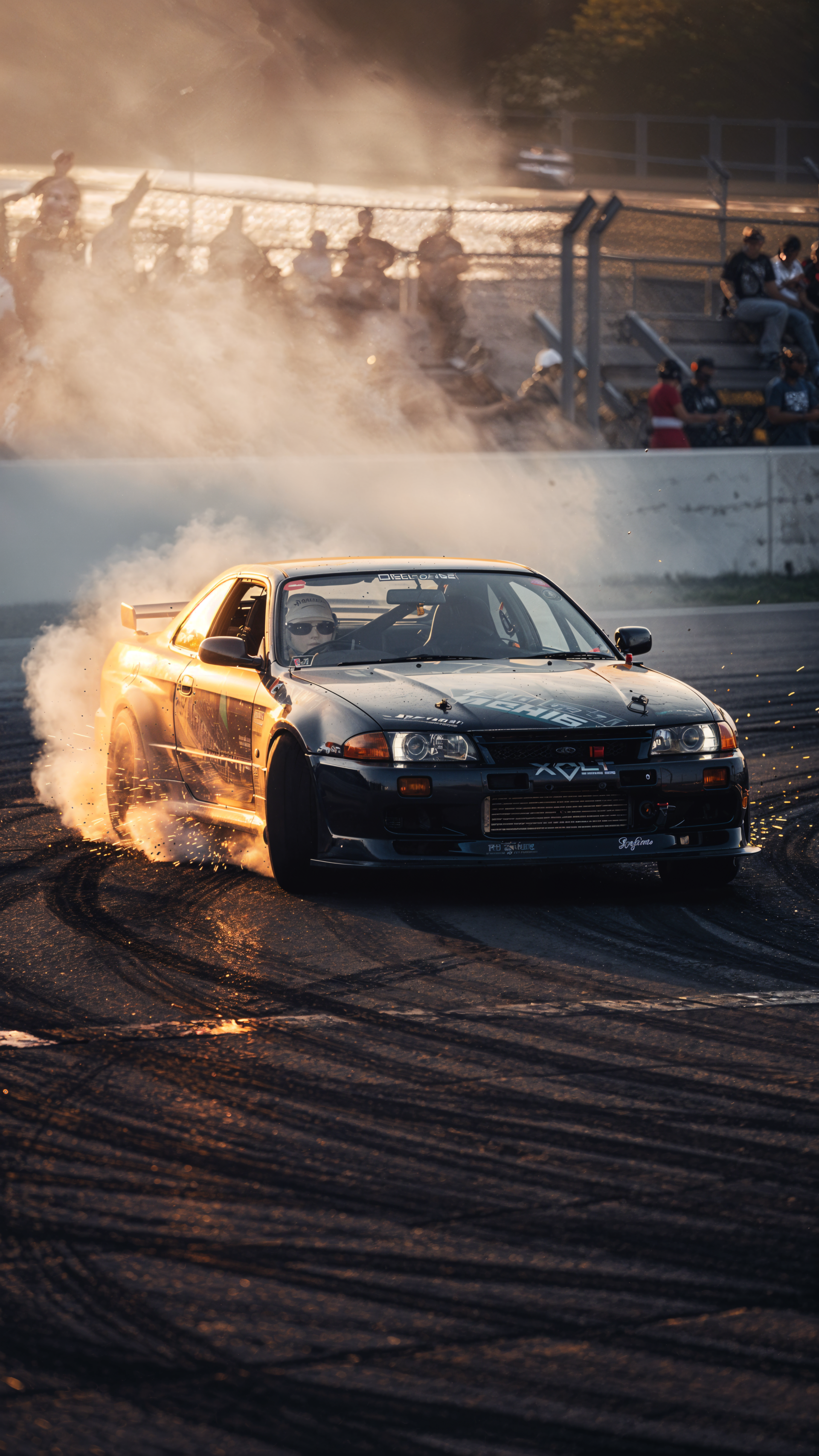 Drifting phone wallpaper: a sports car sliding through a sunlit racetrack corner, rear tires smoking and carving skid marks, spectators blurred behind safety barriers.