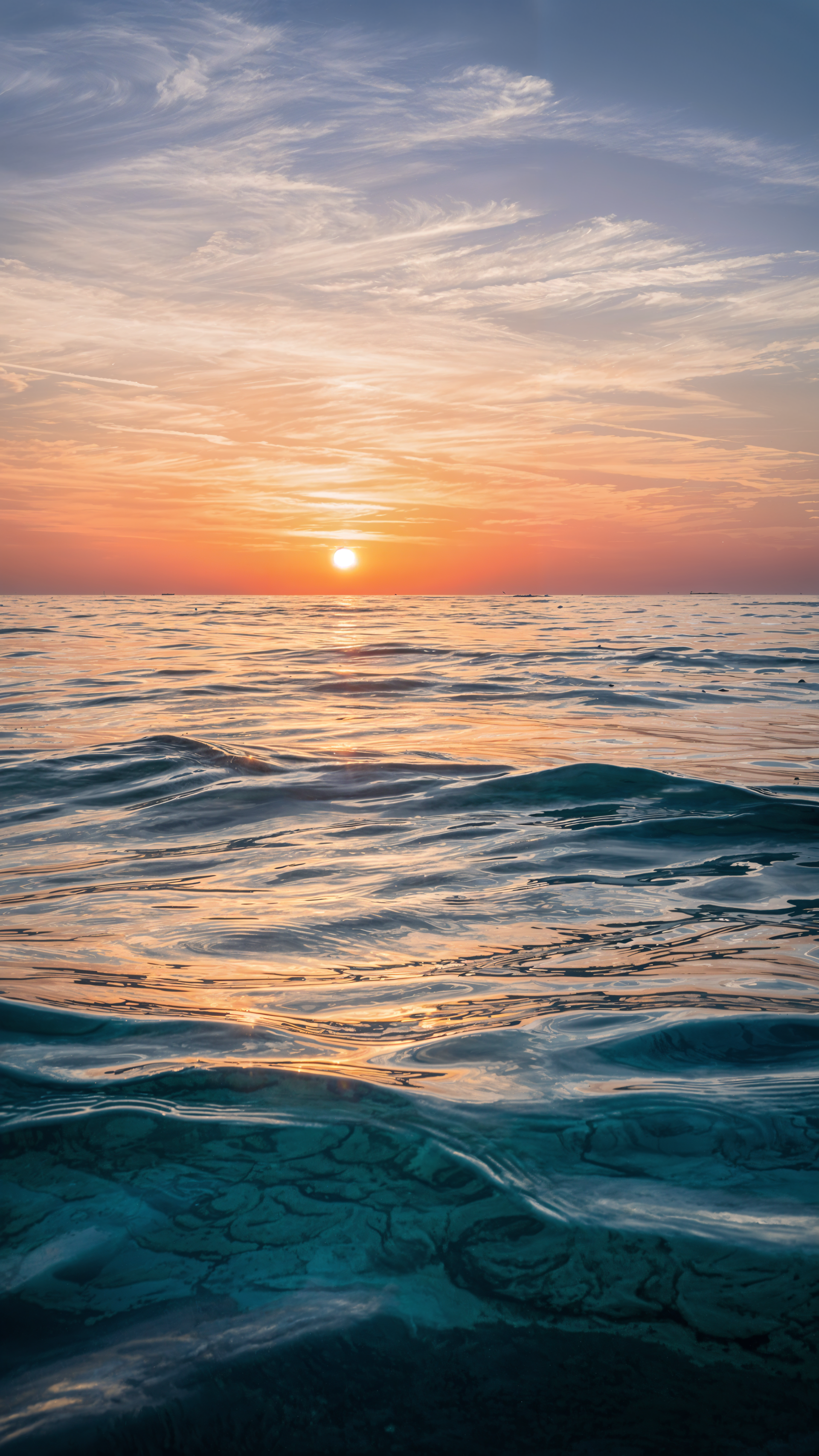 Vertical iPhone wallpaper showing ocean horizon at sunset — turquoise ripples in the foreground, a glowing sun near the skyline and soft pastel clouds above.