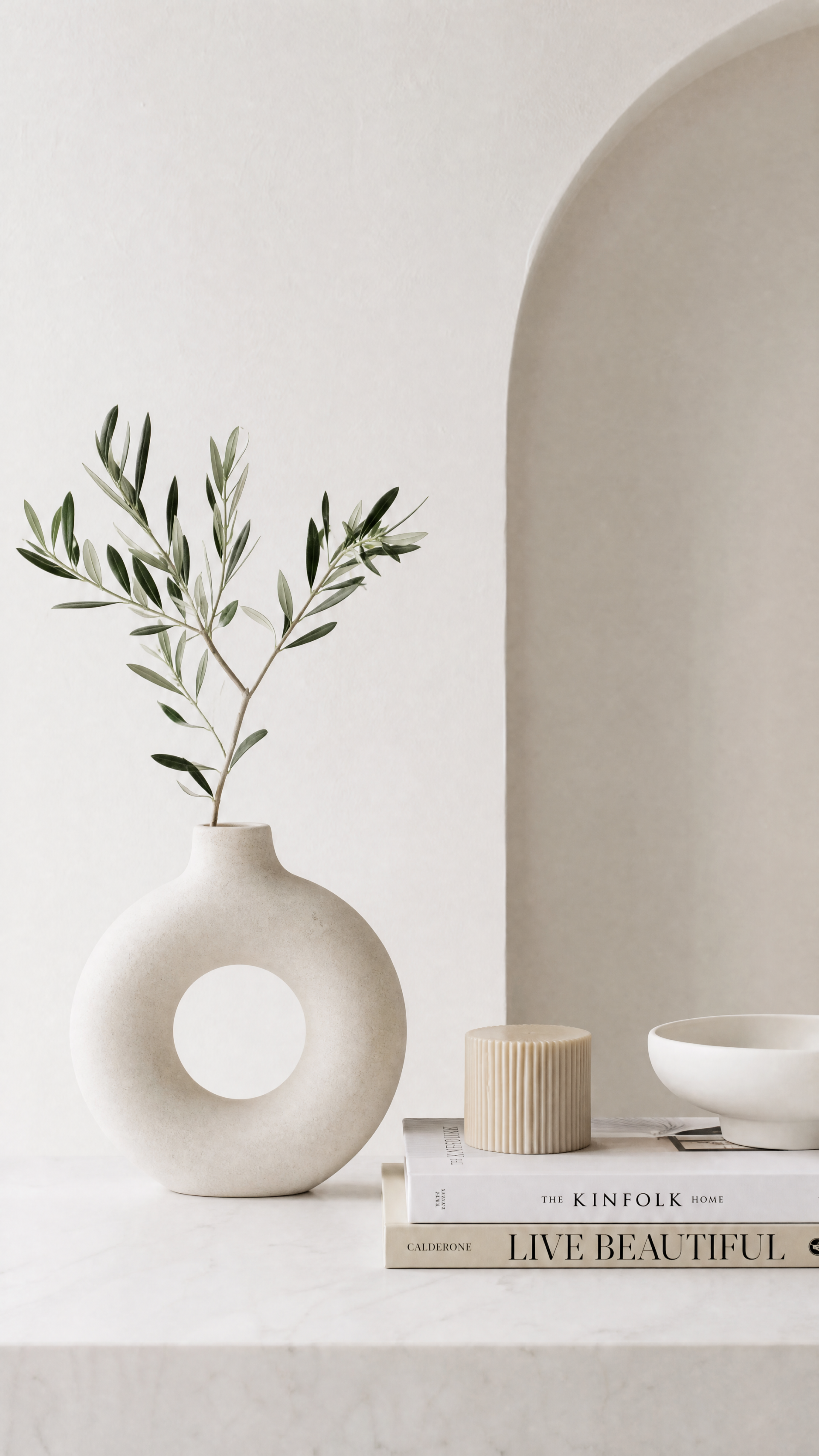 Clean iPhone wallpaper — minimalist still life with a round white vase holding olive branches, stacked neutral books, a small bowl, and a soft arched niche on a beige background.
