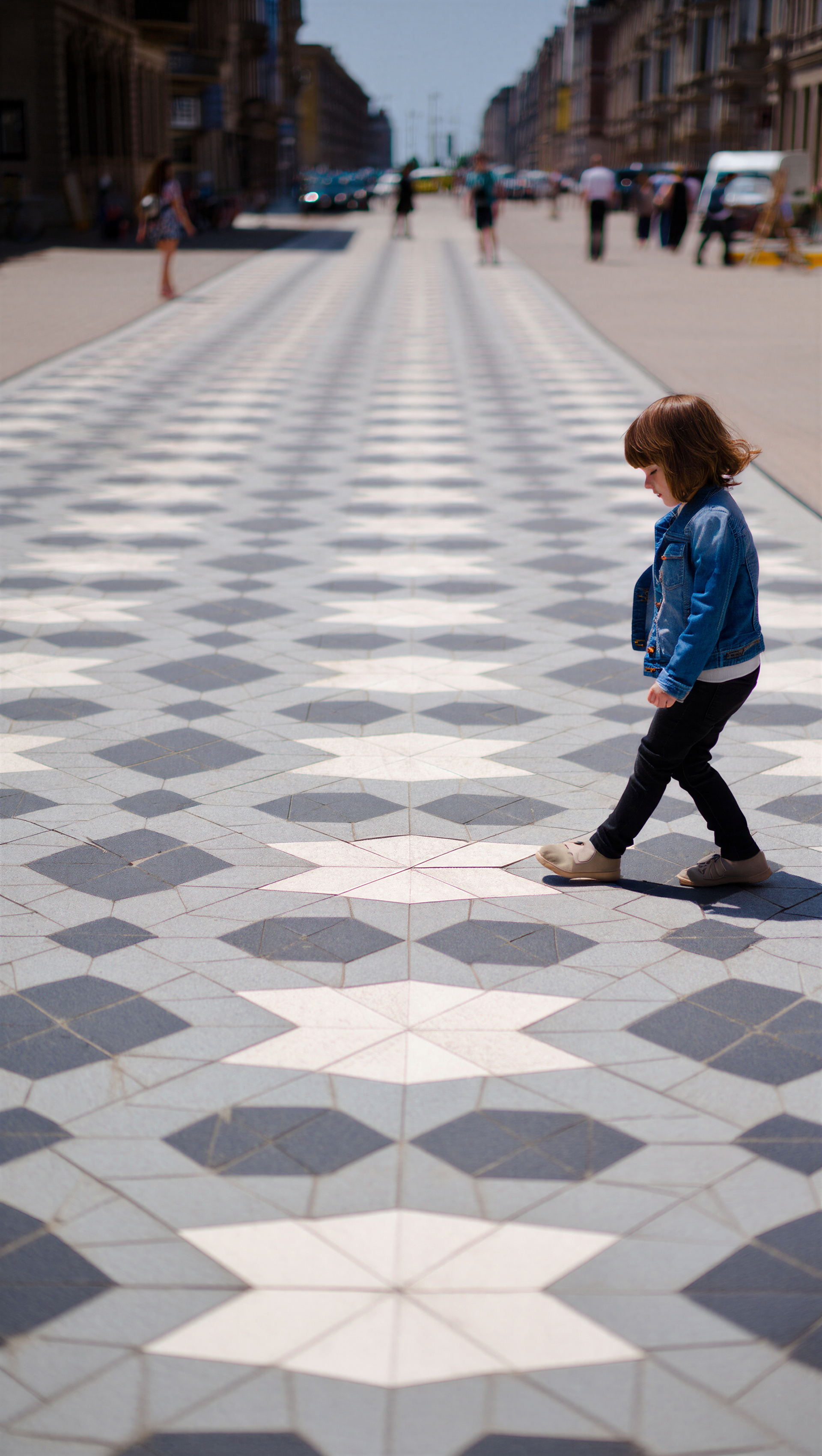Street photography iPhone wallpaper: a child in a blue jacket walks across a geometric tiled plaza, deep perspective with blurred figures and city buildings receding into the distance.