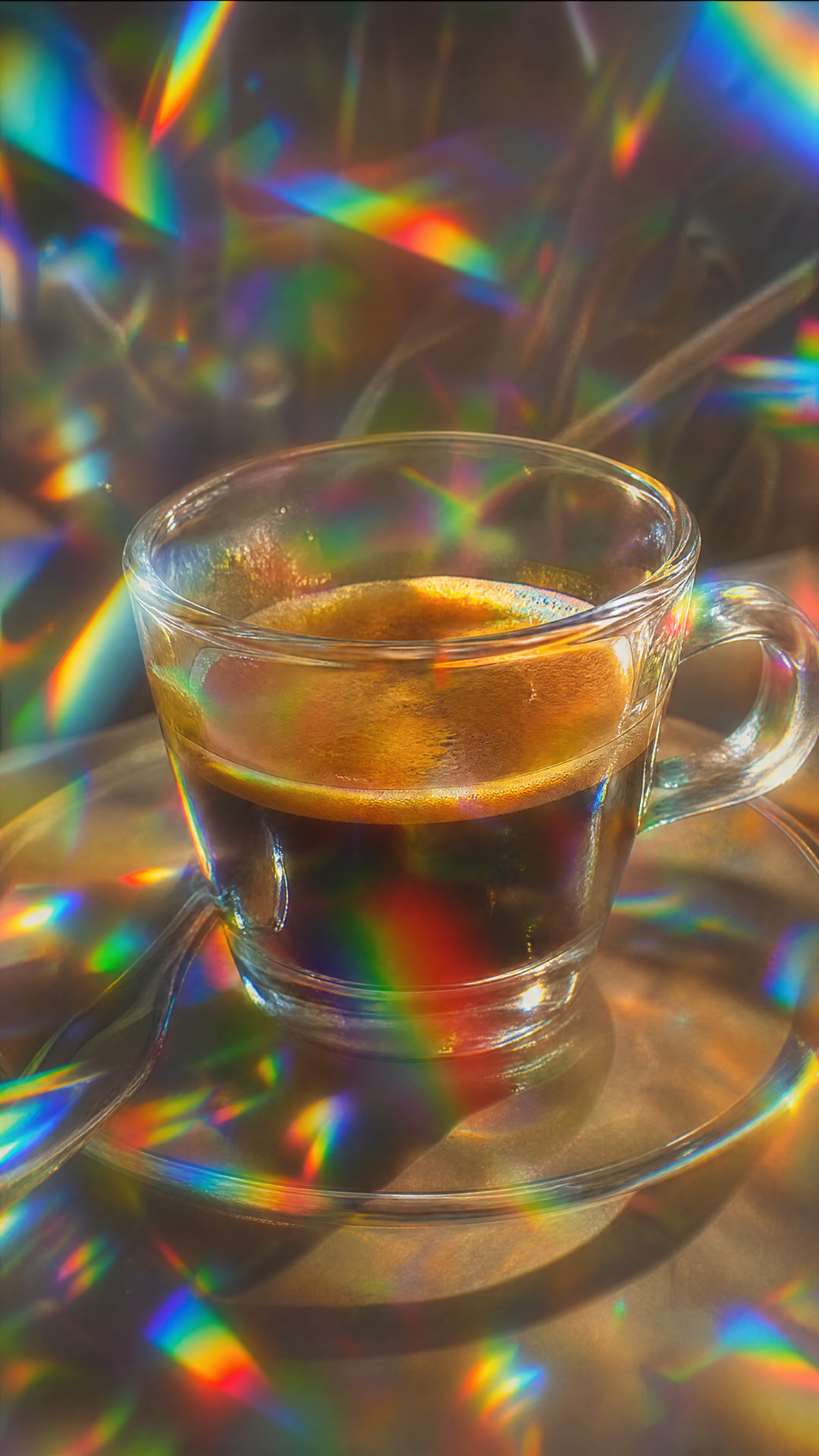 Glass cup of espresso on a saucer, bathed in prismatic rainbow light — vertical phone/iPhone wallpaper.