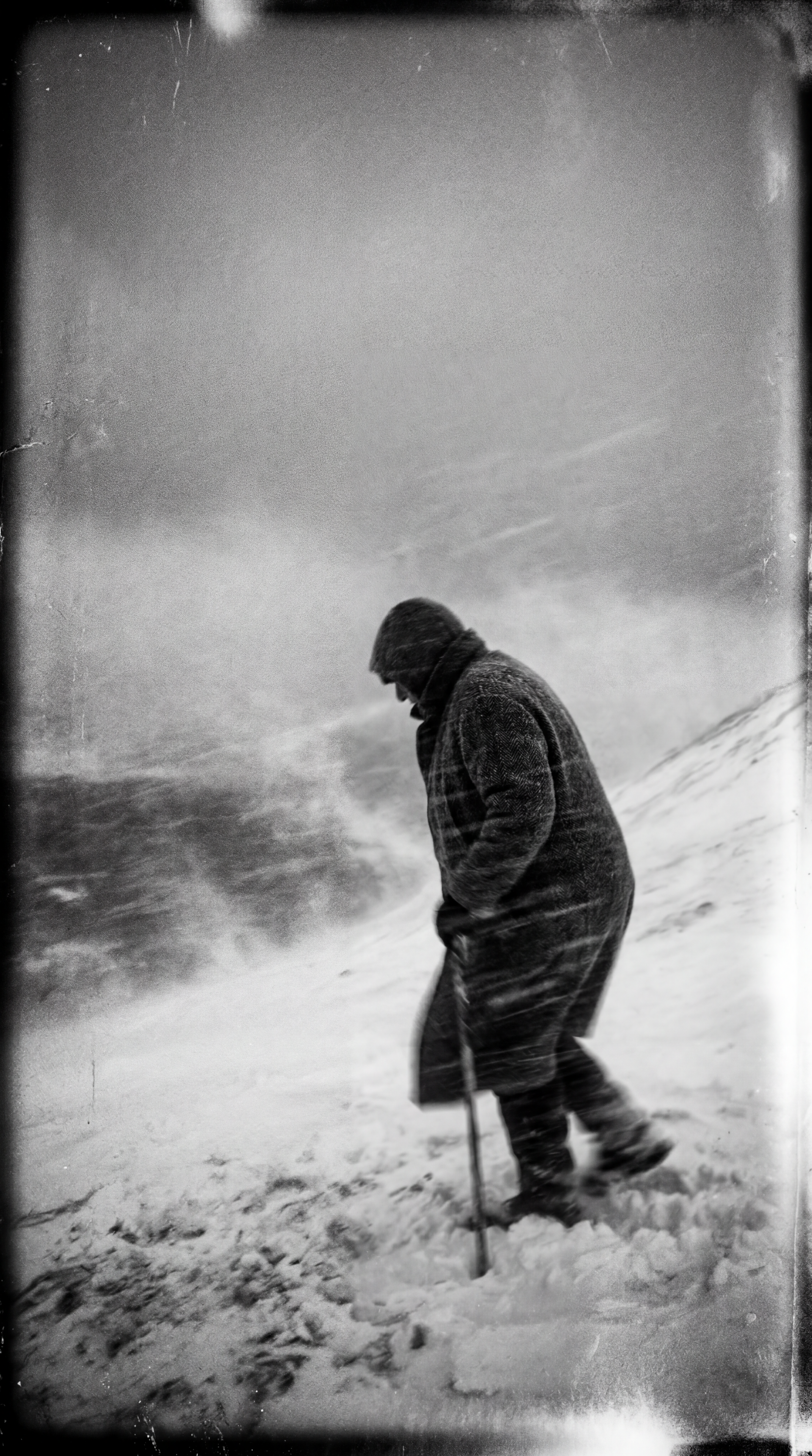 Black-and-white iPhone wallpaper showing a lone figure trudging through a blizzard, hunched against the wind with a walking stick, snow and grainy texture filling a stark, desolate landscape.