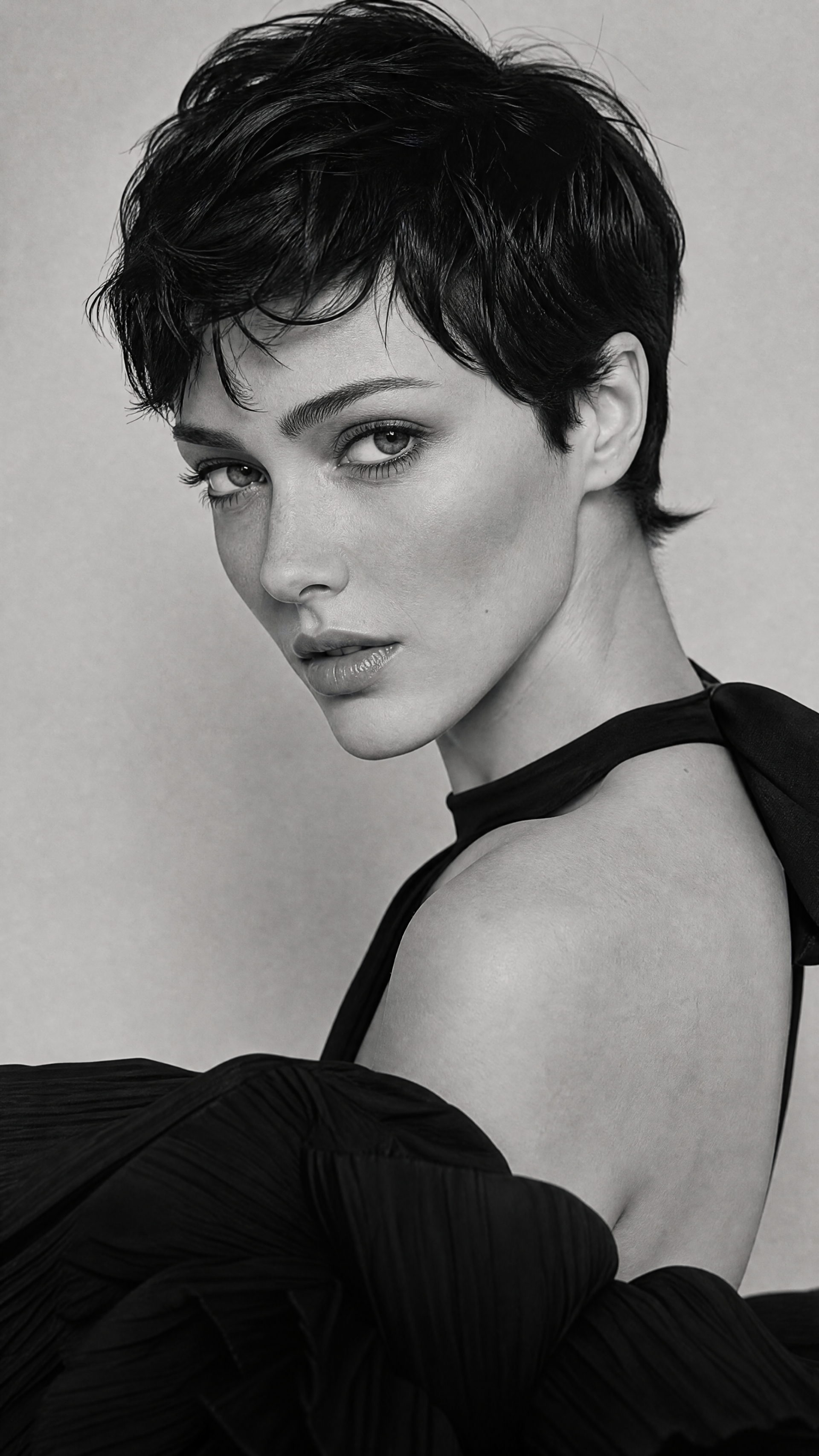 Monochrome close-up of a woman with short dark hair, turned over her shoulder and meeting the camera with an intense gaze; sleek minimalist iPhone wallpaper.