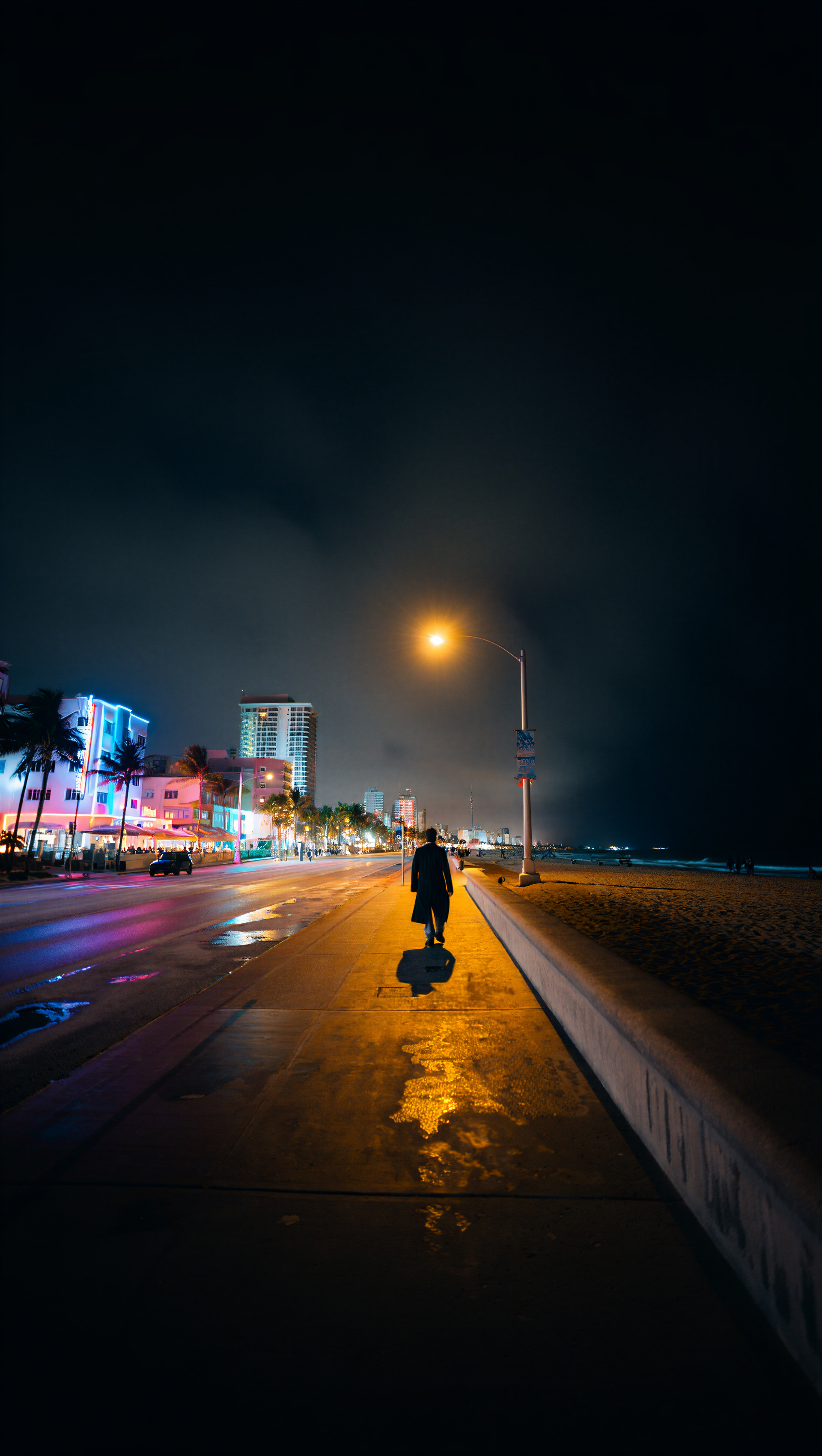 Vertical iPhone wallpaper: lone figure walking a wet Miami beachfront promenade at night, neon-lit buildings and a streetlamp reflecting on the pavement.