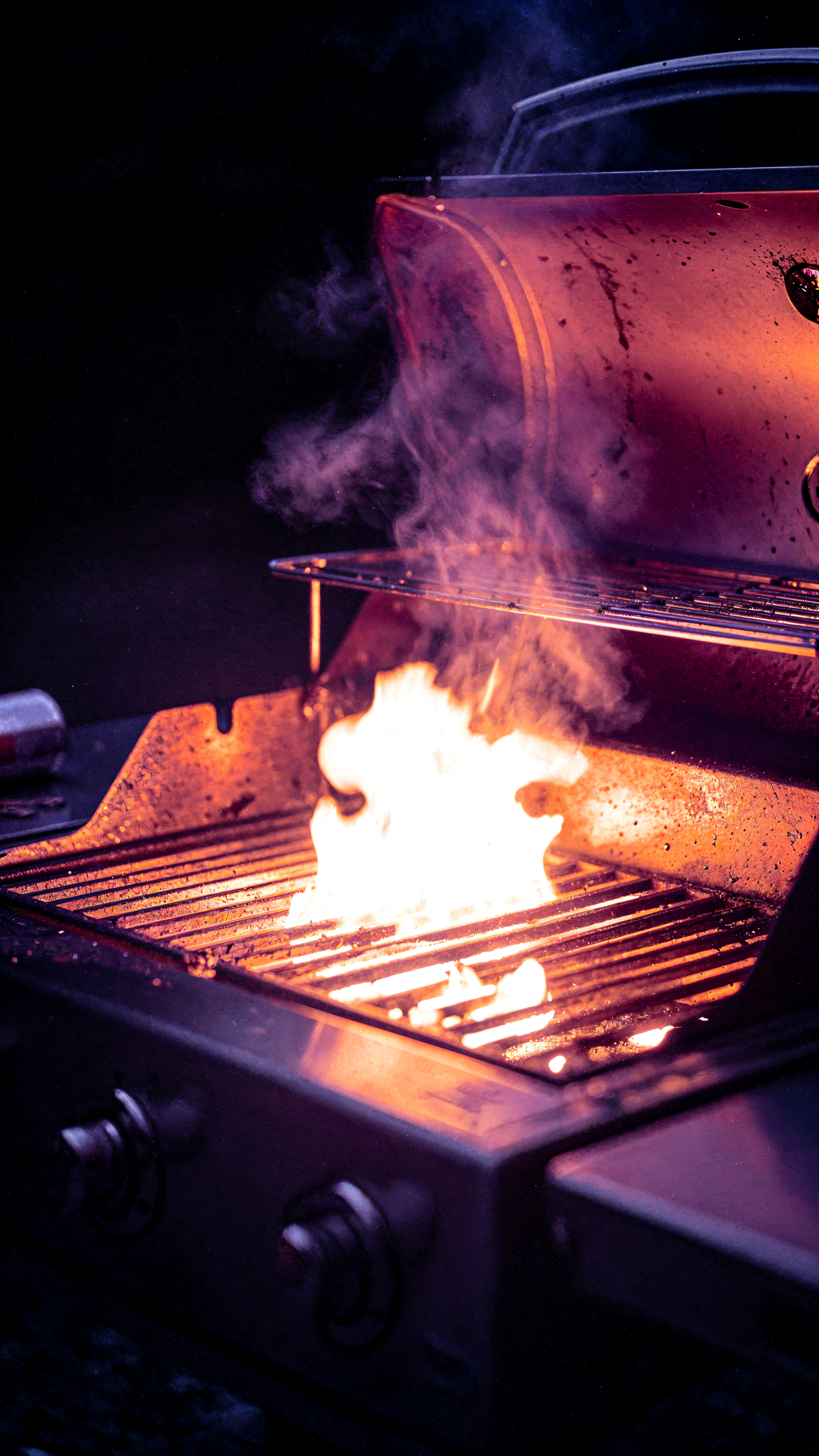 iPhone wallpaper showing a close-up grill with bright flames and curling smoke over hot metal grates, warm orange glow against a dark background.