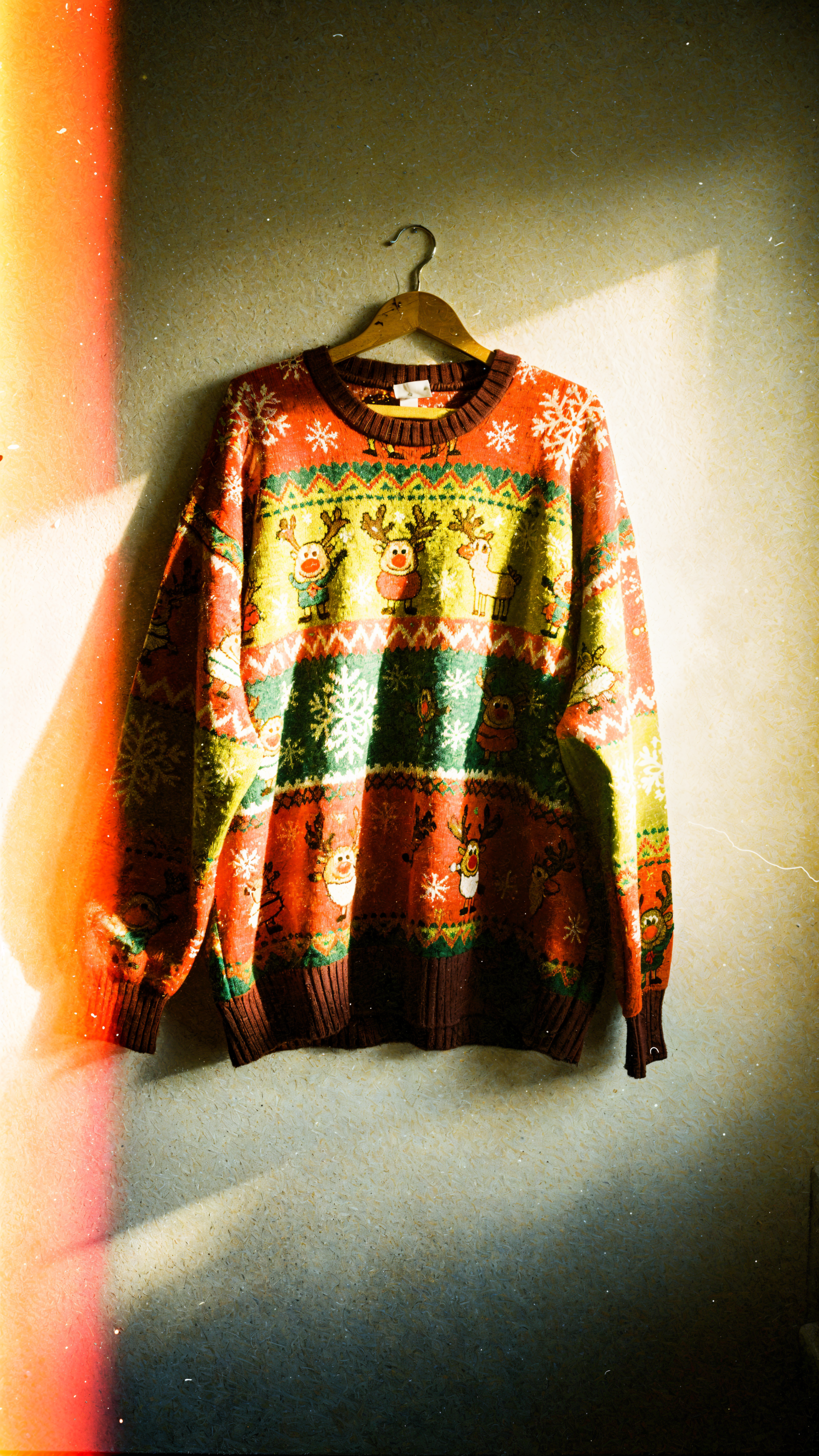 iPhone wallpaper: an ugly red-green Christmas sweater with reindeer and snowflakes hanging on a wooden hanger against a textured wall.