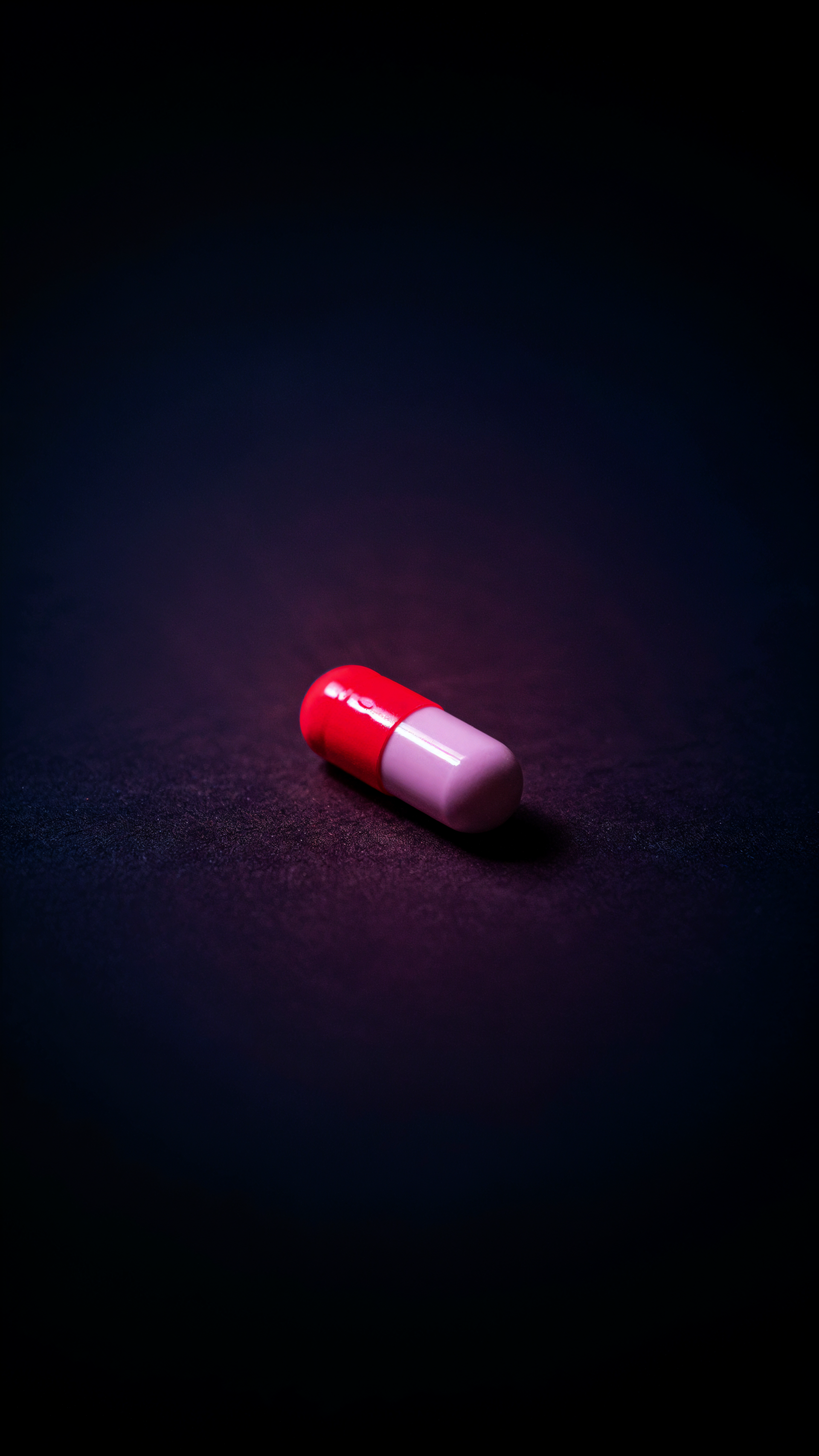iPhone wallpaper showing a single red-and-white capsule pill resting on a dark, moody background with soft spotlighting.