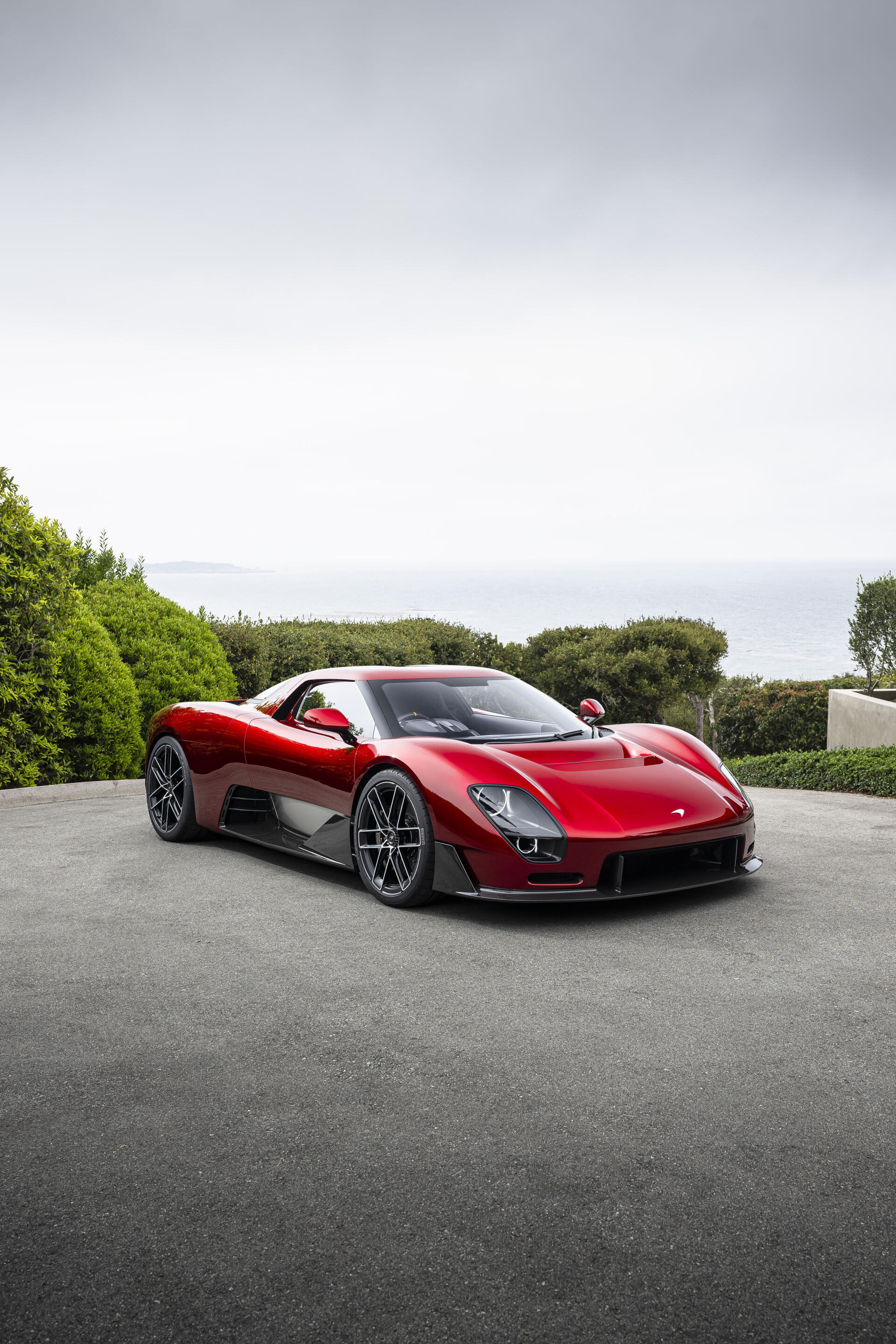 Portrait iPhone wallpaper of a red McLaren McL 6GT sports car parked on a coastal driveway, framed by greenery and sea in the background.