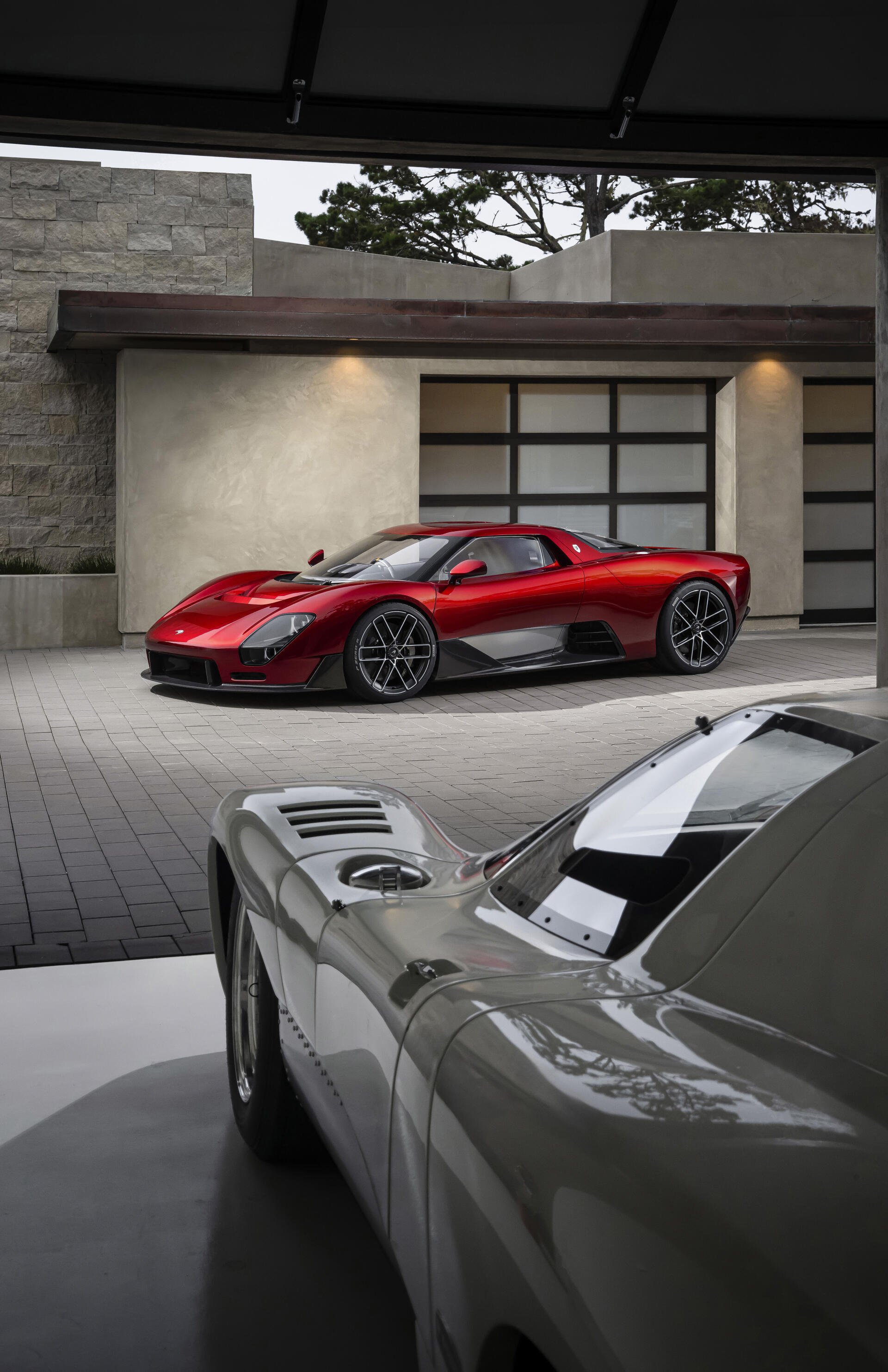 Vertical iPhone wallpaper of a red McLaren McL 6GT supercar parked by a modern garage, another classic sports car vehicle in soft-focus foreground.