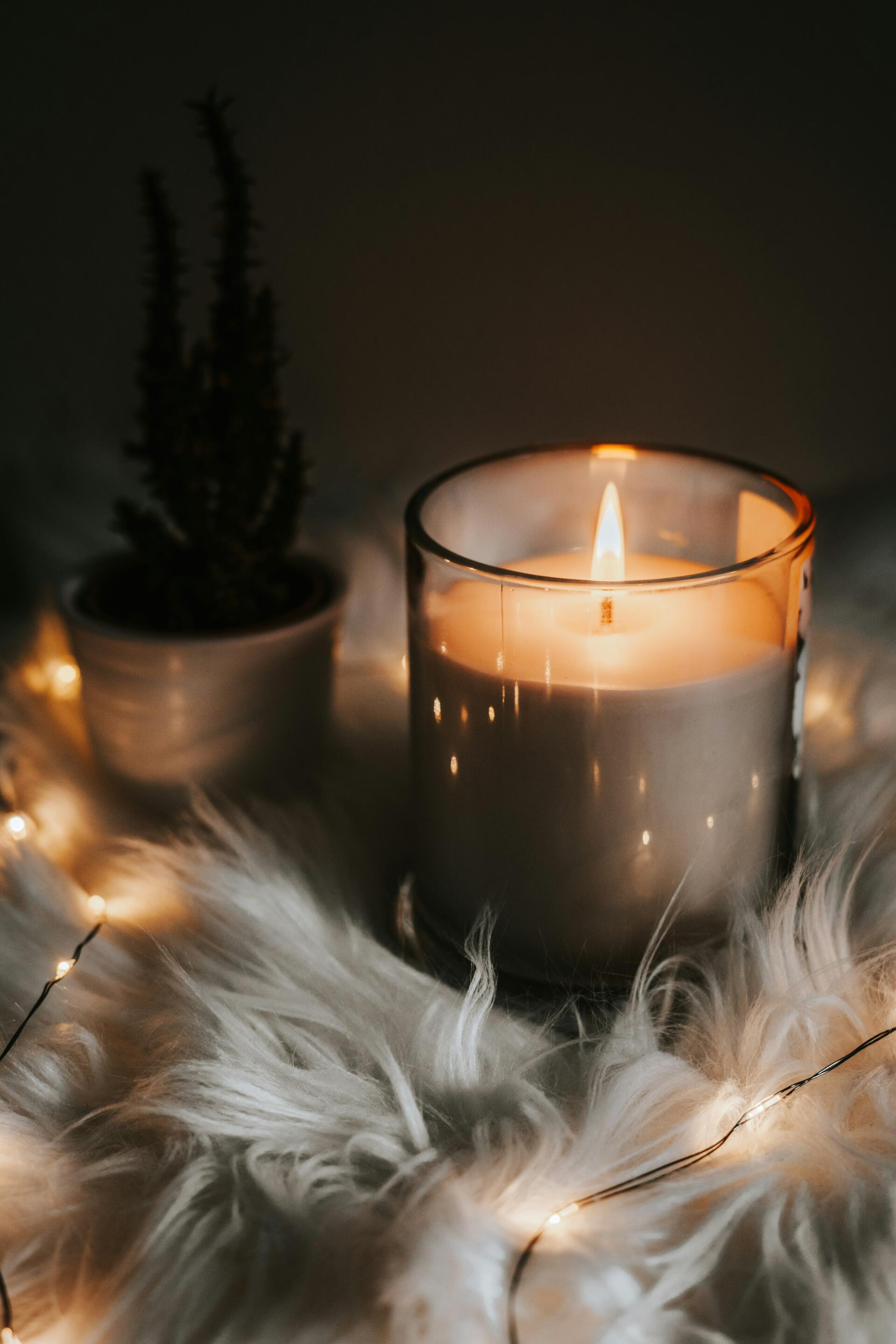 iPhone wallpaper of a lit candle in a glass jar on faux fur with fairy lights and a potted plant, radiating a cozy Kitchen Witch aesthetic.