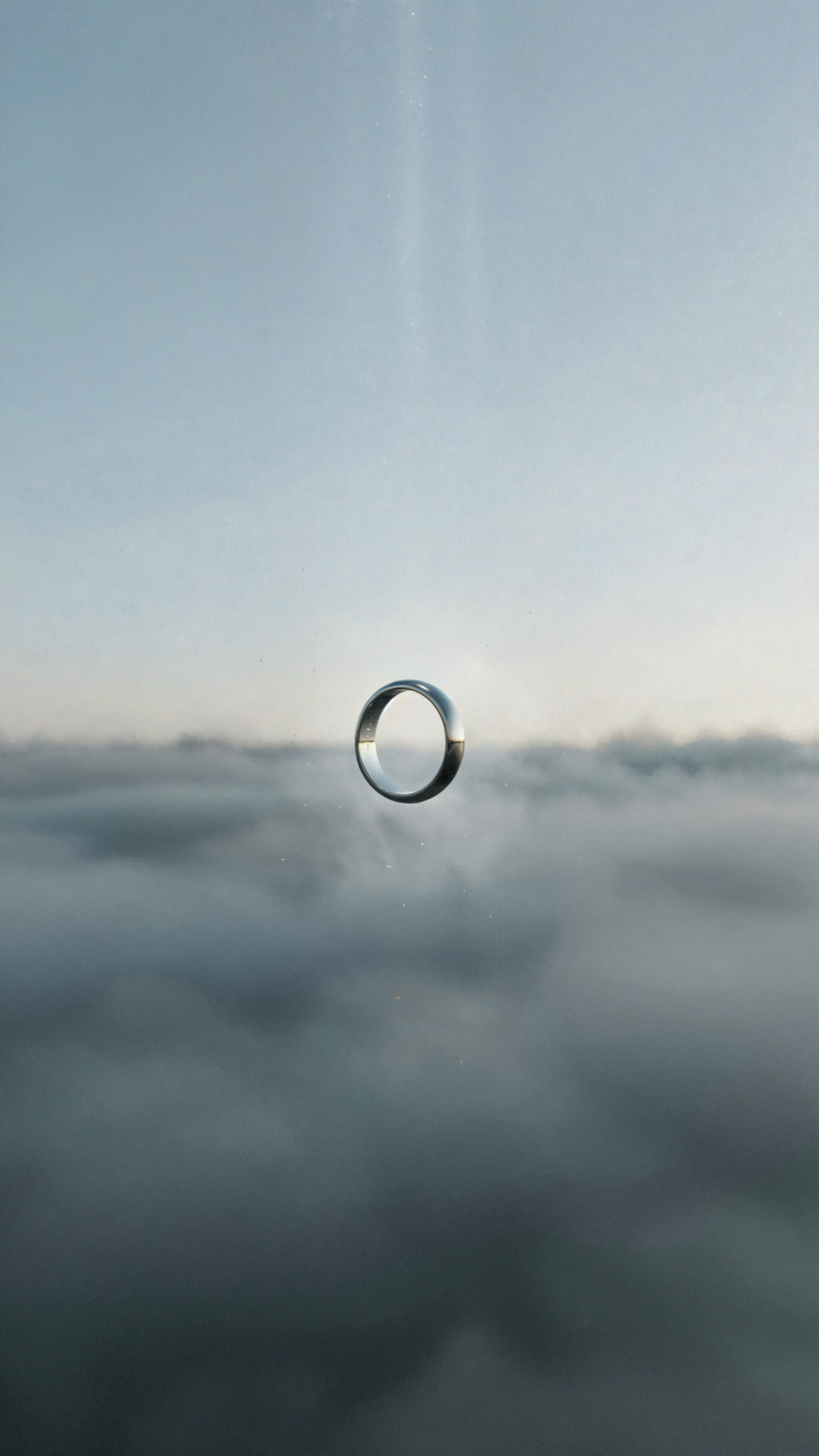 iPhone wallpaper showing a wedding ring suspended like a droplet above misty clouds under a pale sky with a faint vertical beam of light.