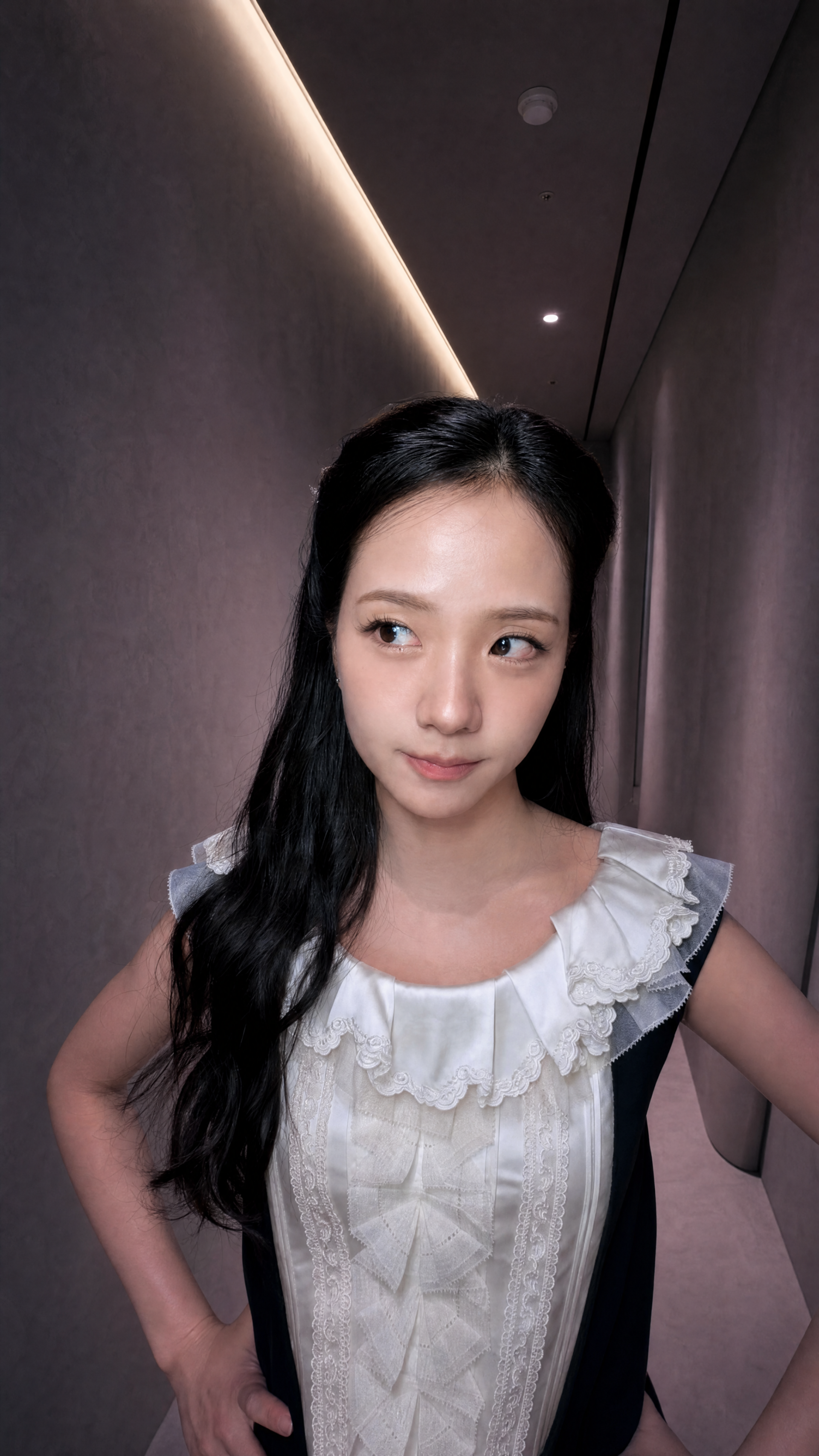 iPhone wallpaper portrait: a young woman with long dark hair in a ruffled white blouse, hands on hips in a narrow, softly lit hallway.
