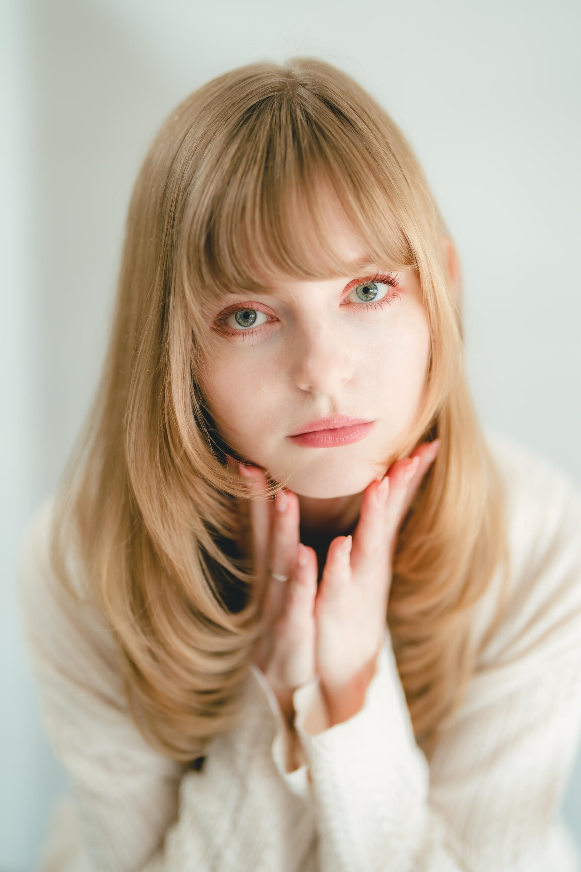 Soft studio photography portrait of a blonde model with green eyes and hands near her chin, minimalist iPhone wallpaper.