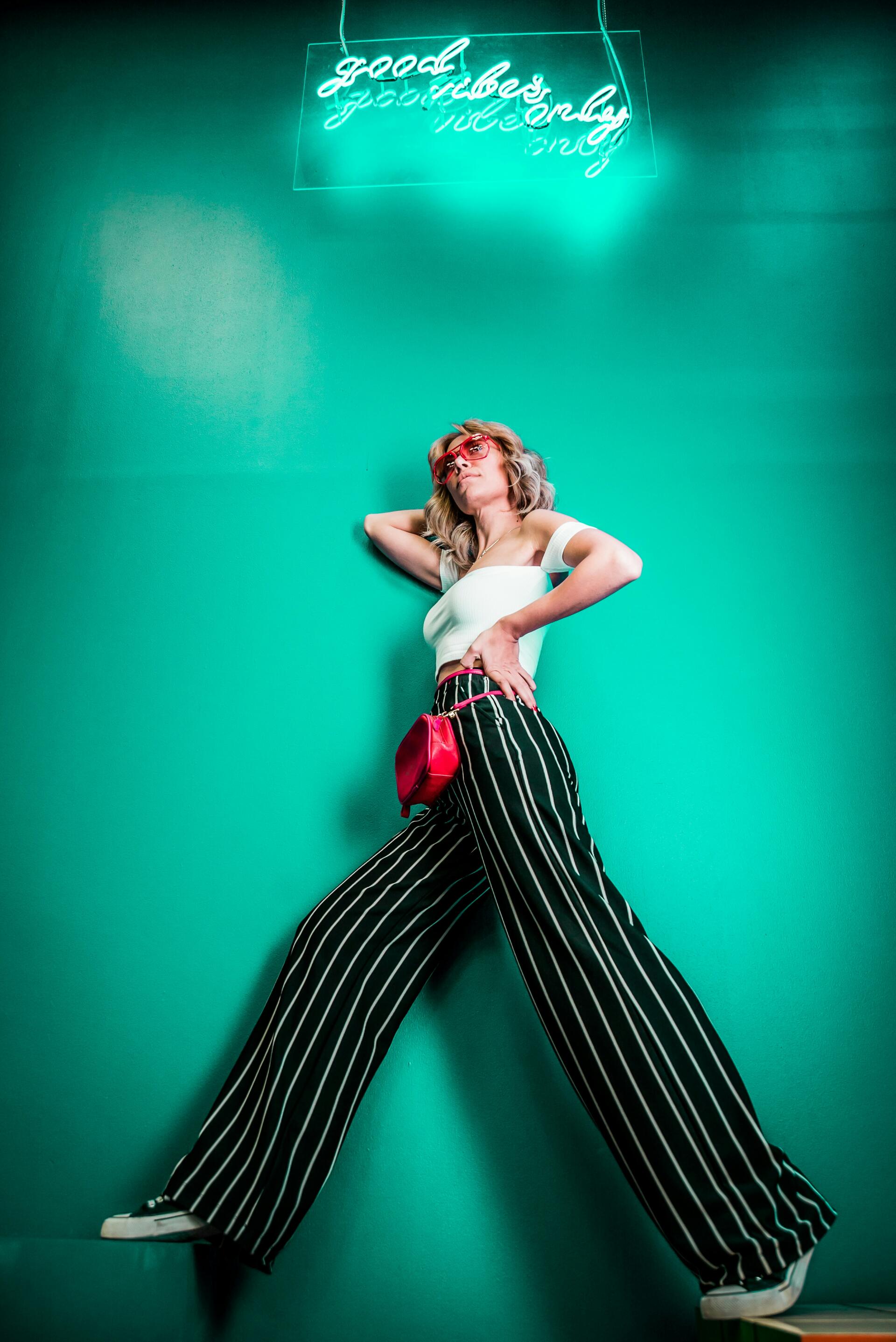 iPhone wallpaper: fashion shot of a woman model in electric yellow accessories, white top and wide striped trousers, posing against a vivid teal wall with neon sign.