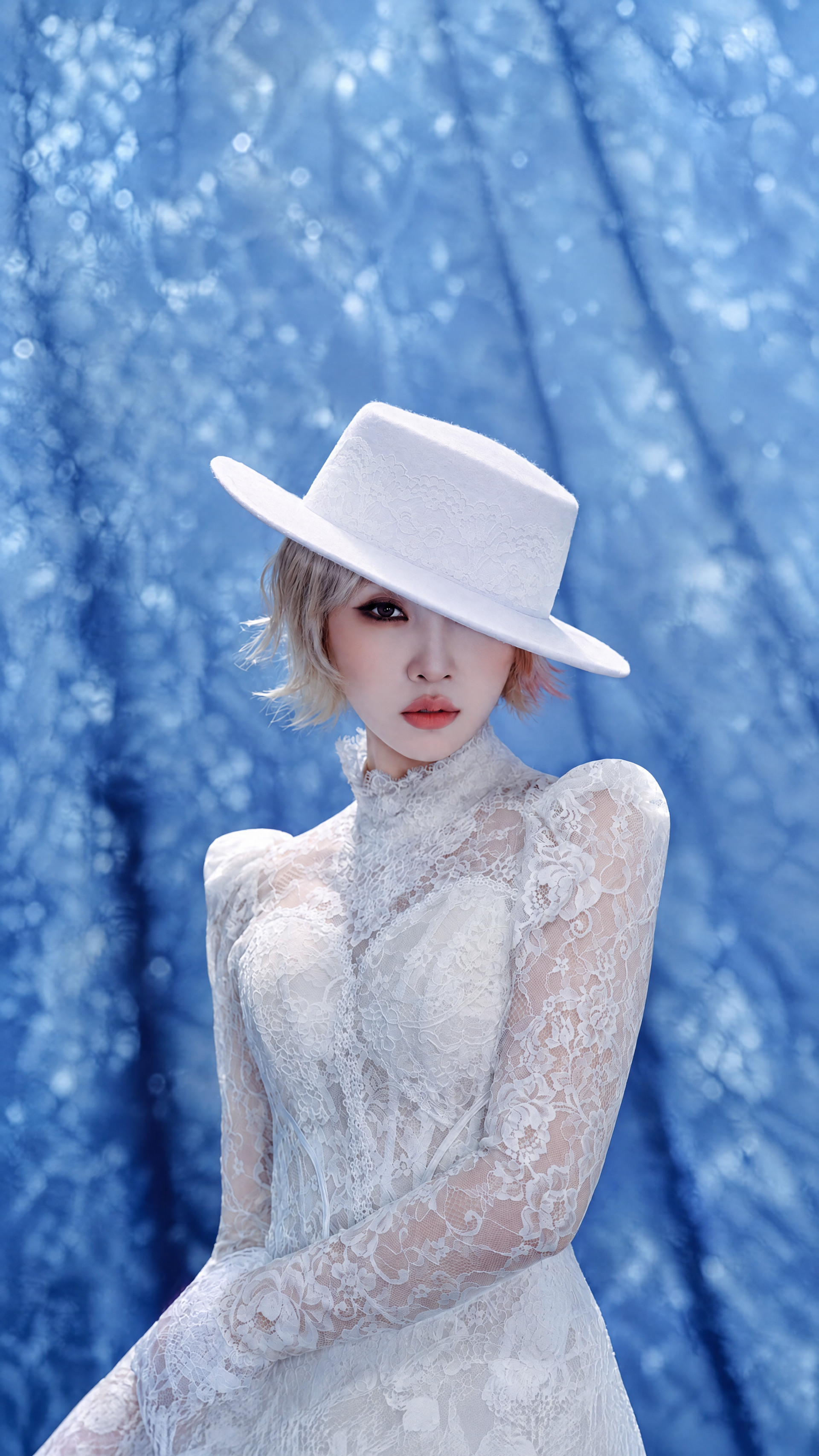 iPhone wallpaper of a woman in a white lace dress and wide-brim white hat posed against a dreamy blue bokeh background, evoking the title CALLING.