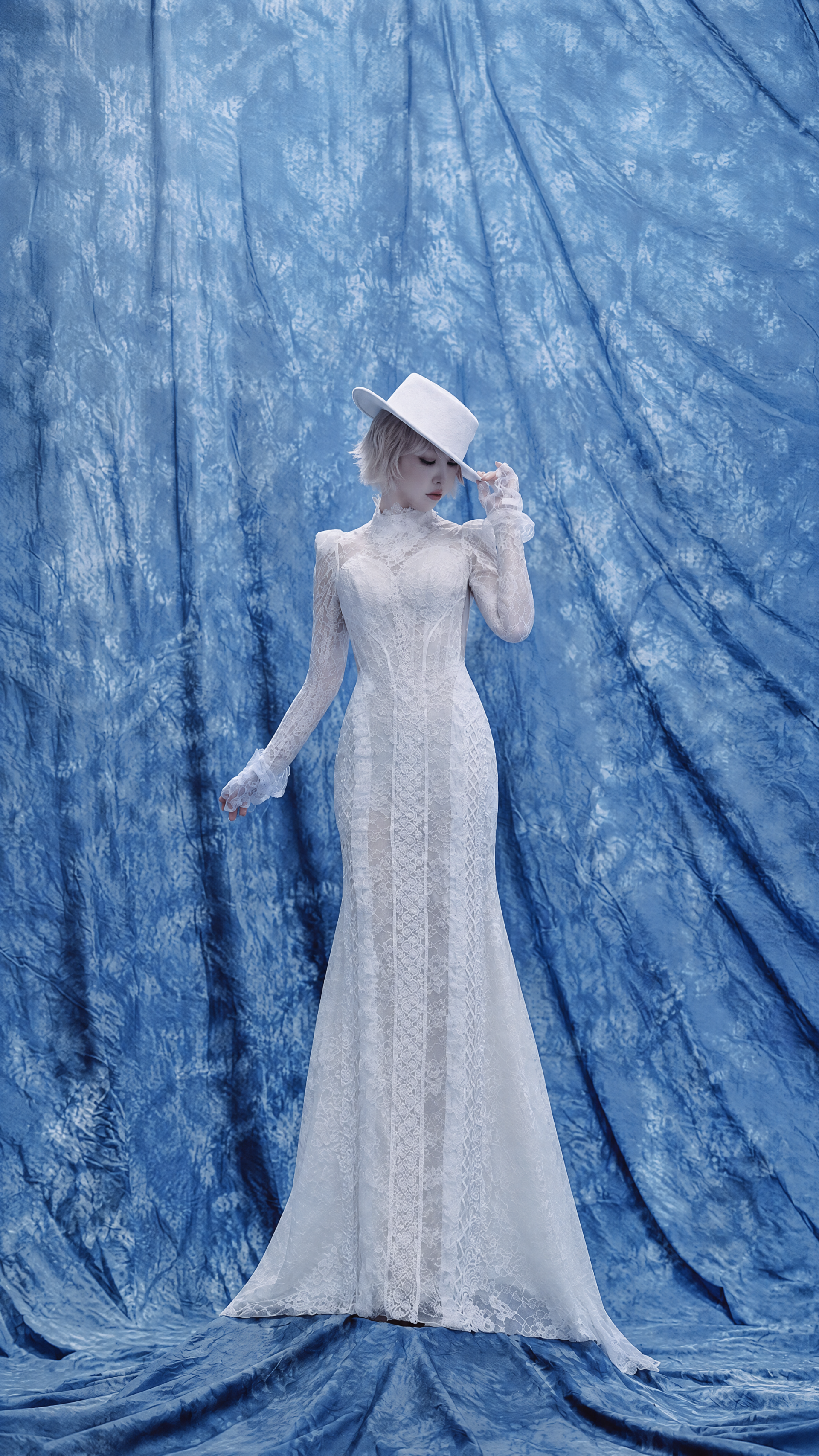 iPhone wallpaper of a woman in a white lace dress and white hat posed against a blue draped backdrop, evoking the theme CALLING.