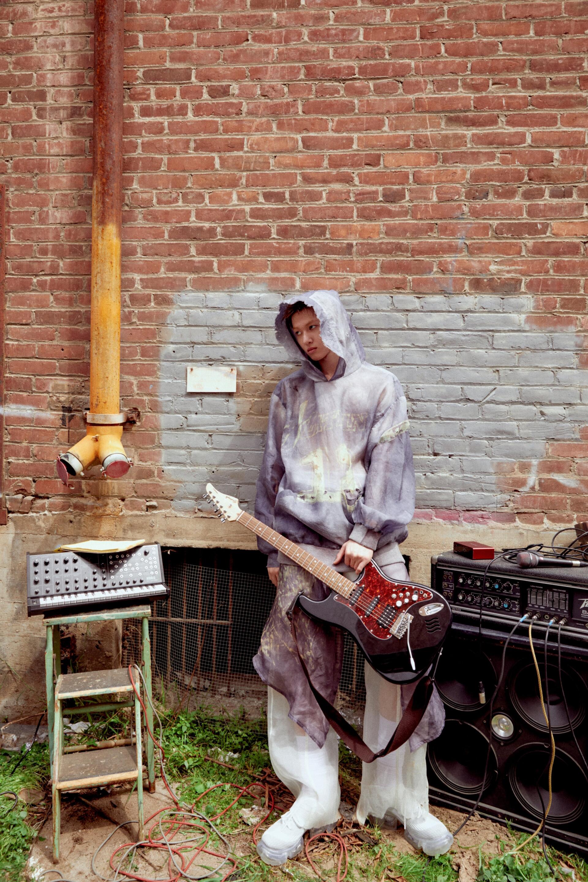 iPhone wallpaper — Cortis, Color Outside the Lines: a hooded figure in paint-splattered clothes holds an electric guitar beside amps against a red-brick wall.