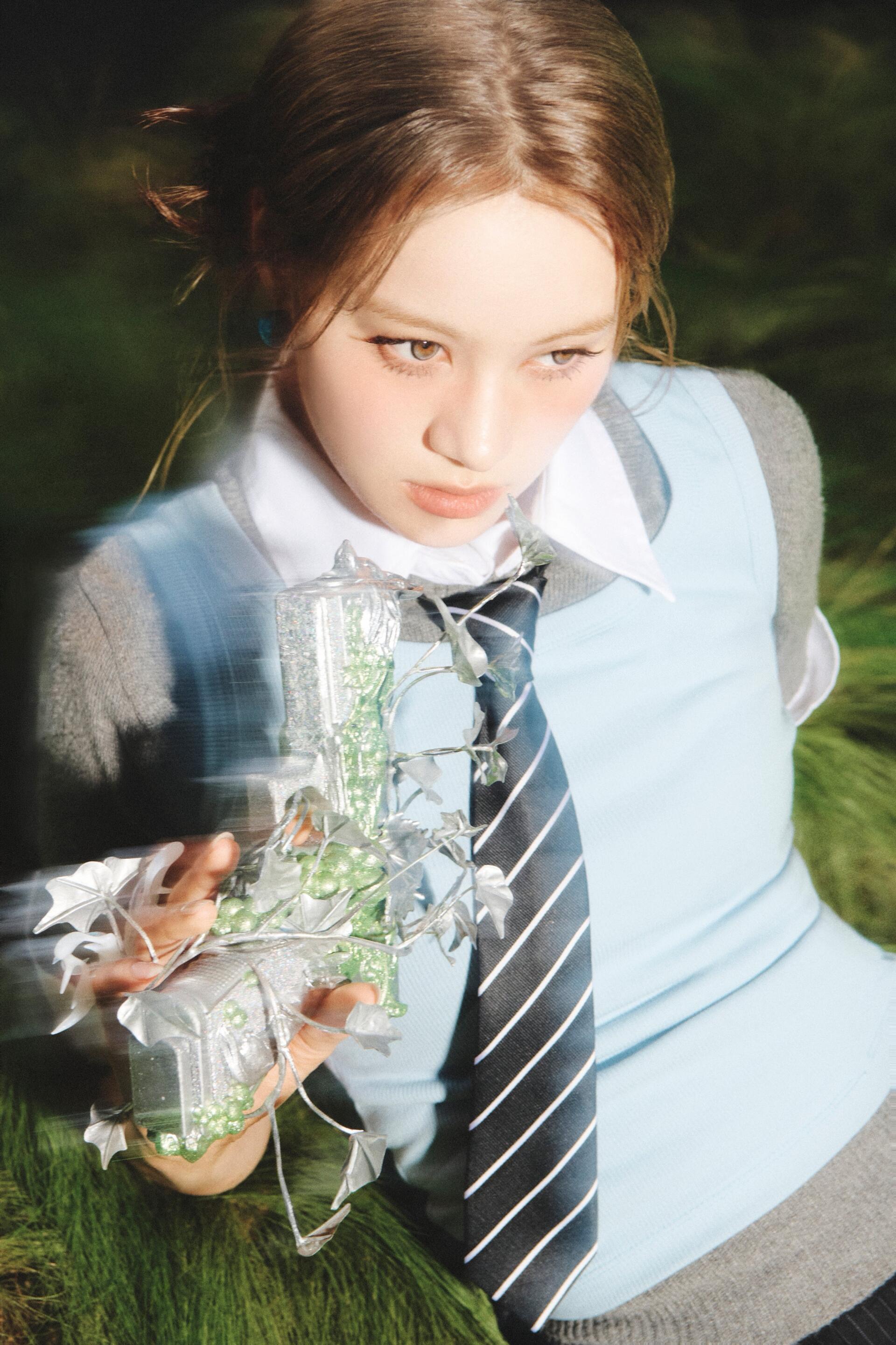 Phone wallpaper of a schoolgirl in uniform and striped tie, reaching with crumpled foil-wrapped green candy — moody 'Sour Grapes' pop-concept vibe.