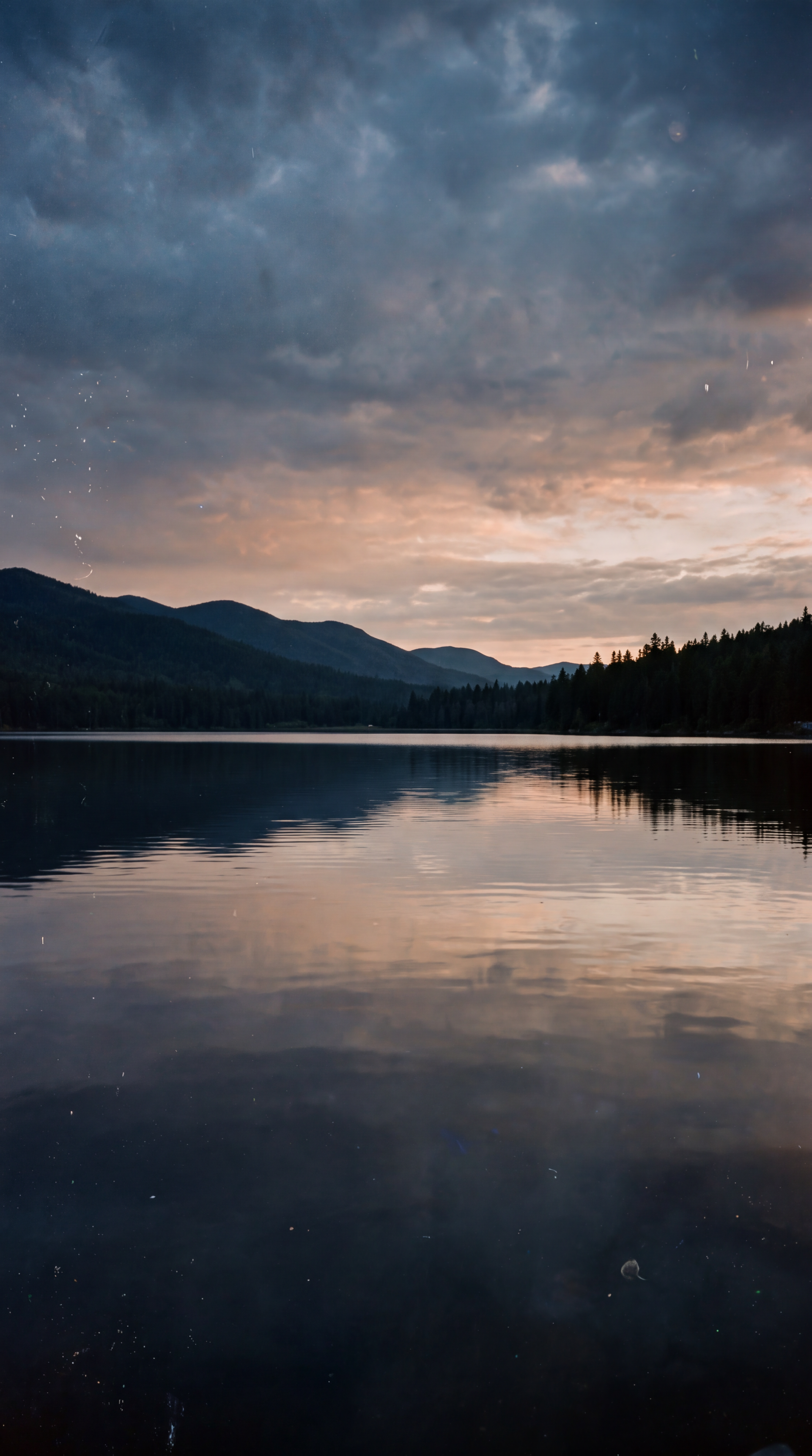 Vertical iPhone wallpaper: tranquil mountain lake at dusk, calm reflective water beneath a cloudy sky.